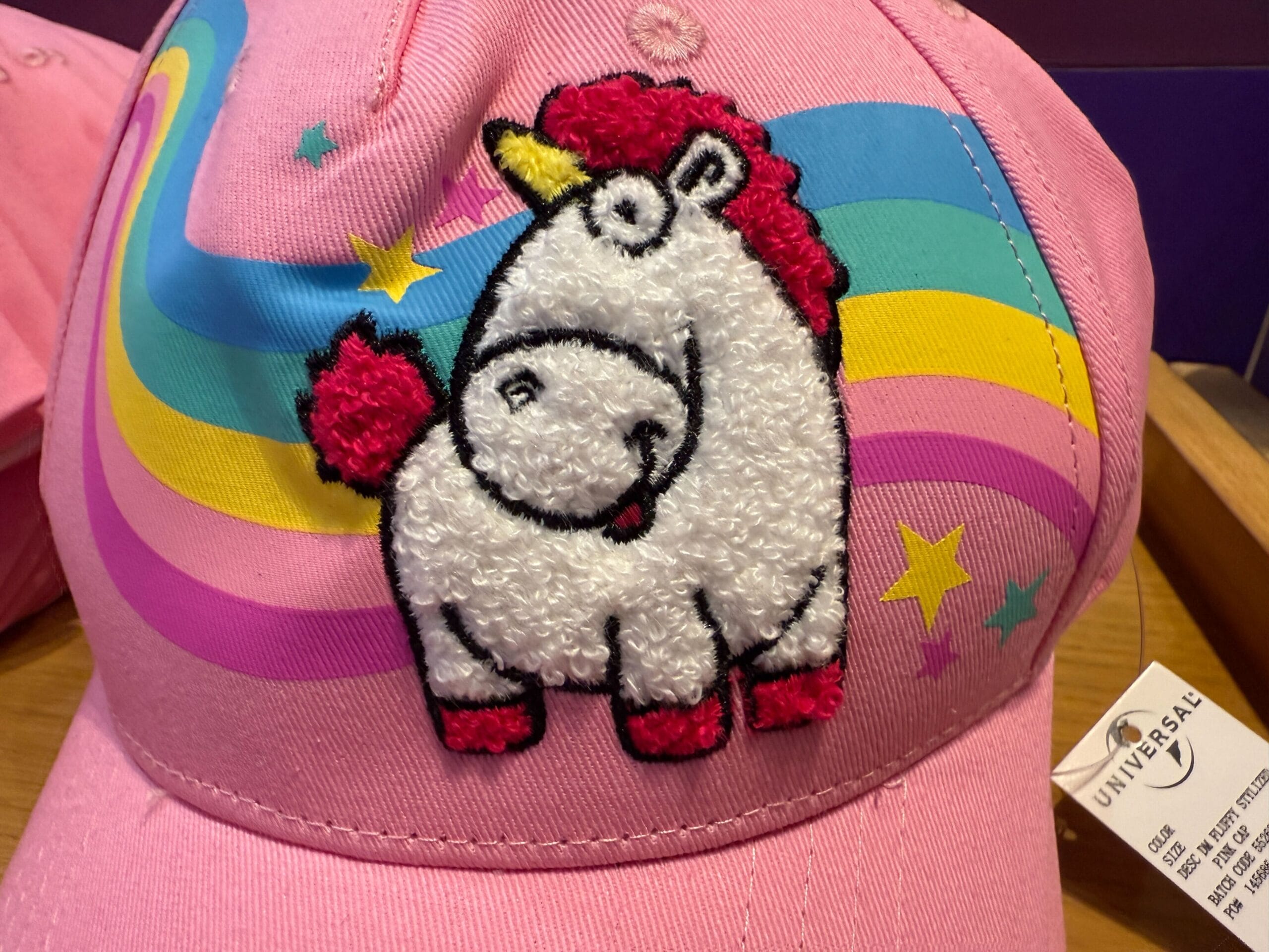 Pink cap with a Fluffy Unicorn patch, rainbow background, yellow stars, and Universal Orlando Resort tag.