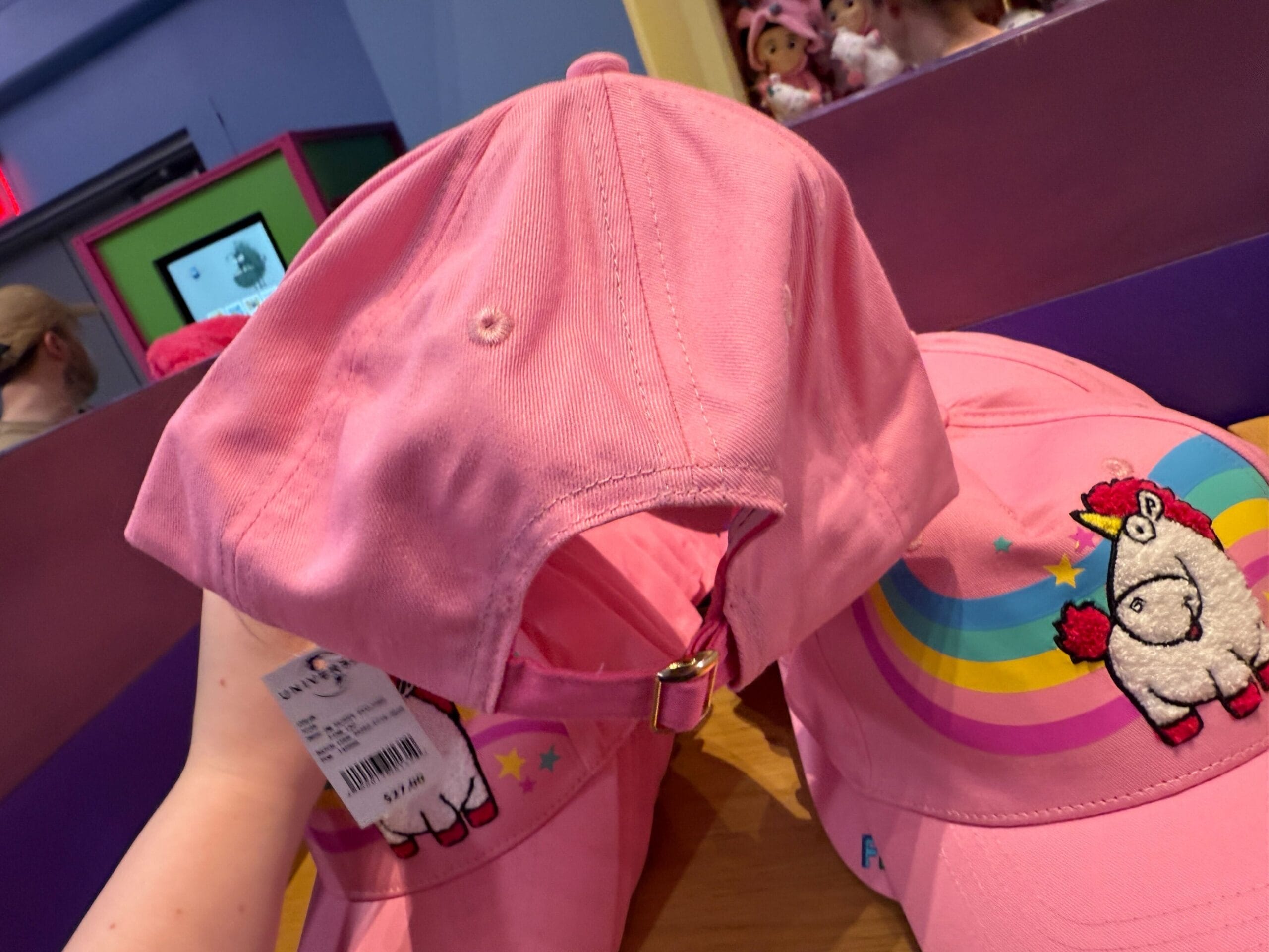 Hand holds a pink cap with a Fluffy Unicorn llama; another cap on the table has rainbow and star designs.