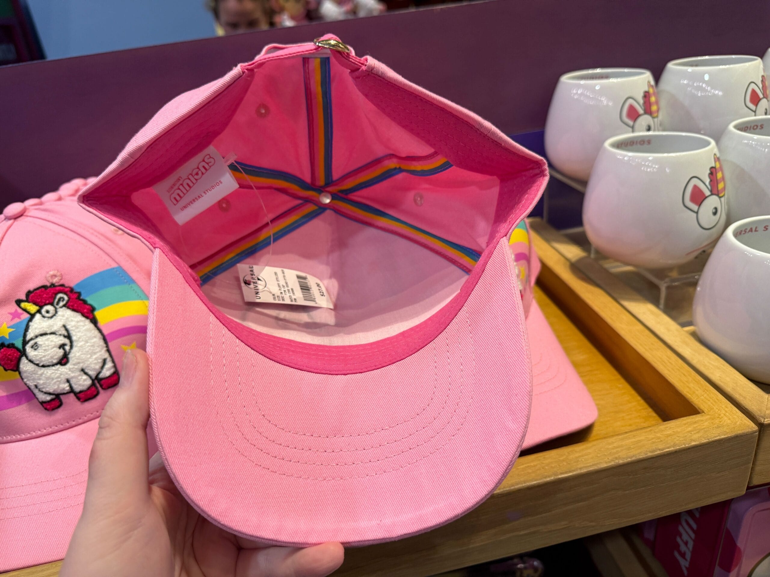 A hand holds a pink cap with rainbow lining, beside Fluffy Unicorn mugs and another unicorn cap at Universal Orlando Resort.