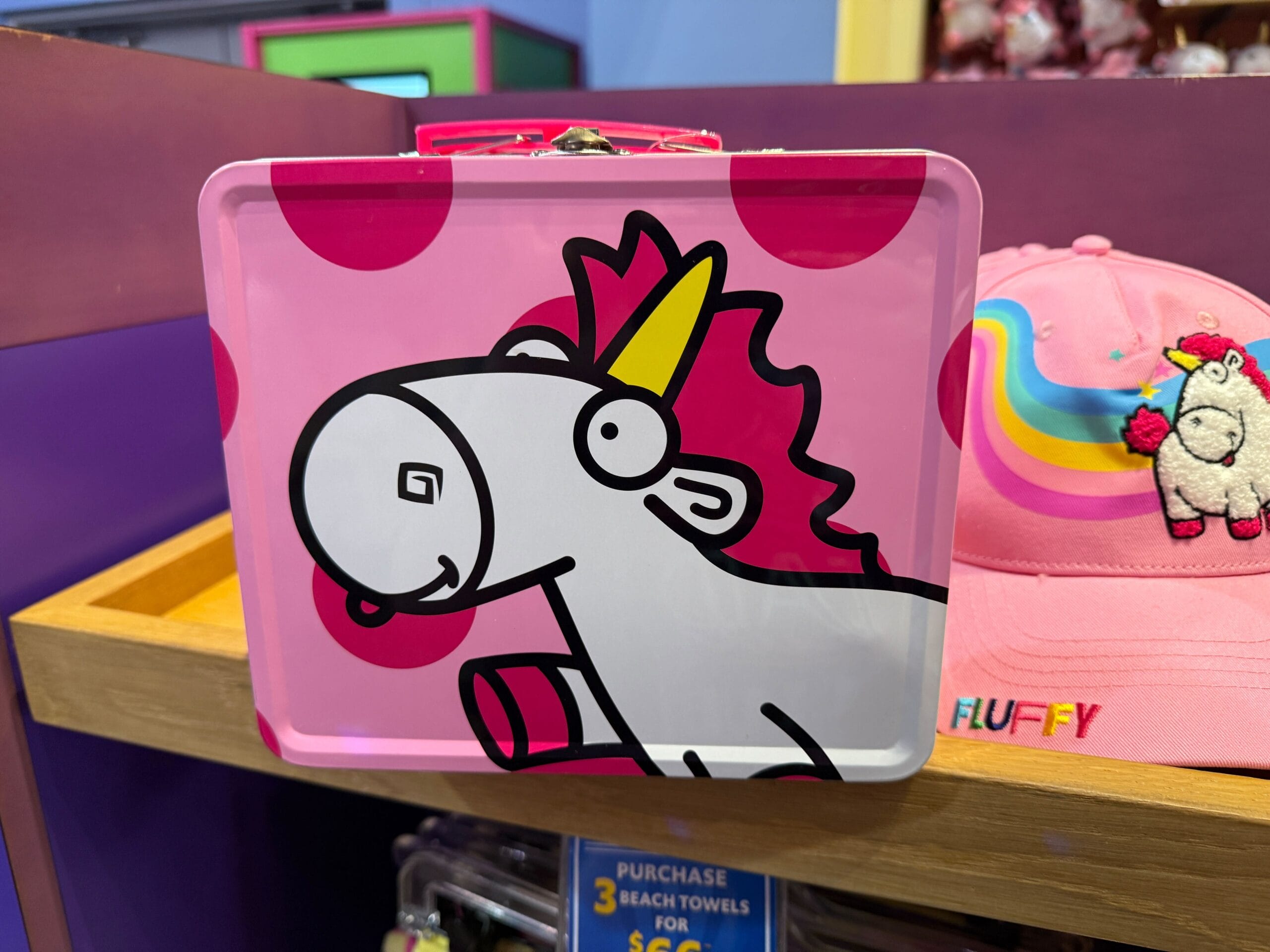 Pink lunchbox with a cartoon unicorn, next to a pink hat featuring a Fluffy Unicorn patch on a wooden shelf.