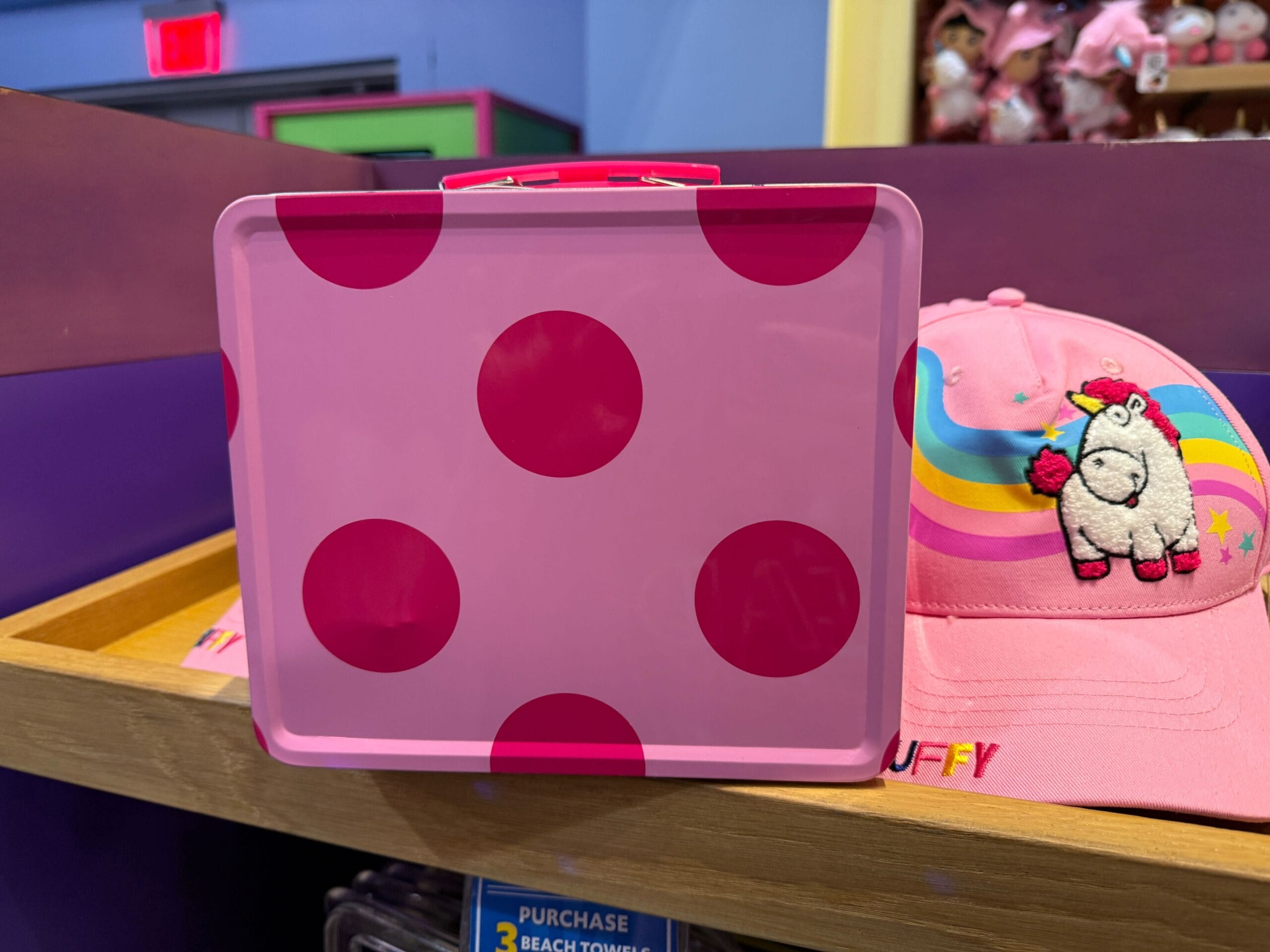 A pink lunchbox with dark pink polka dots sits by a pink Minions hat featuring a Fluffy Unicorn design.