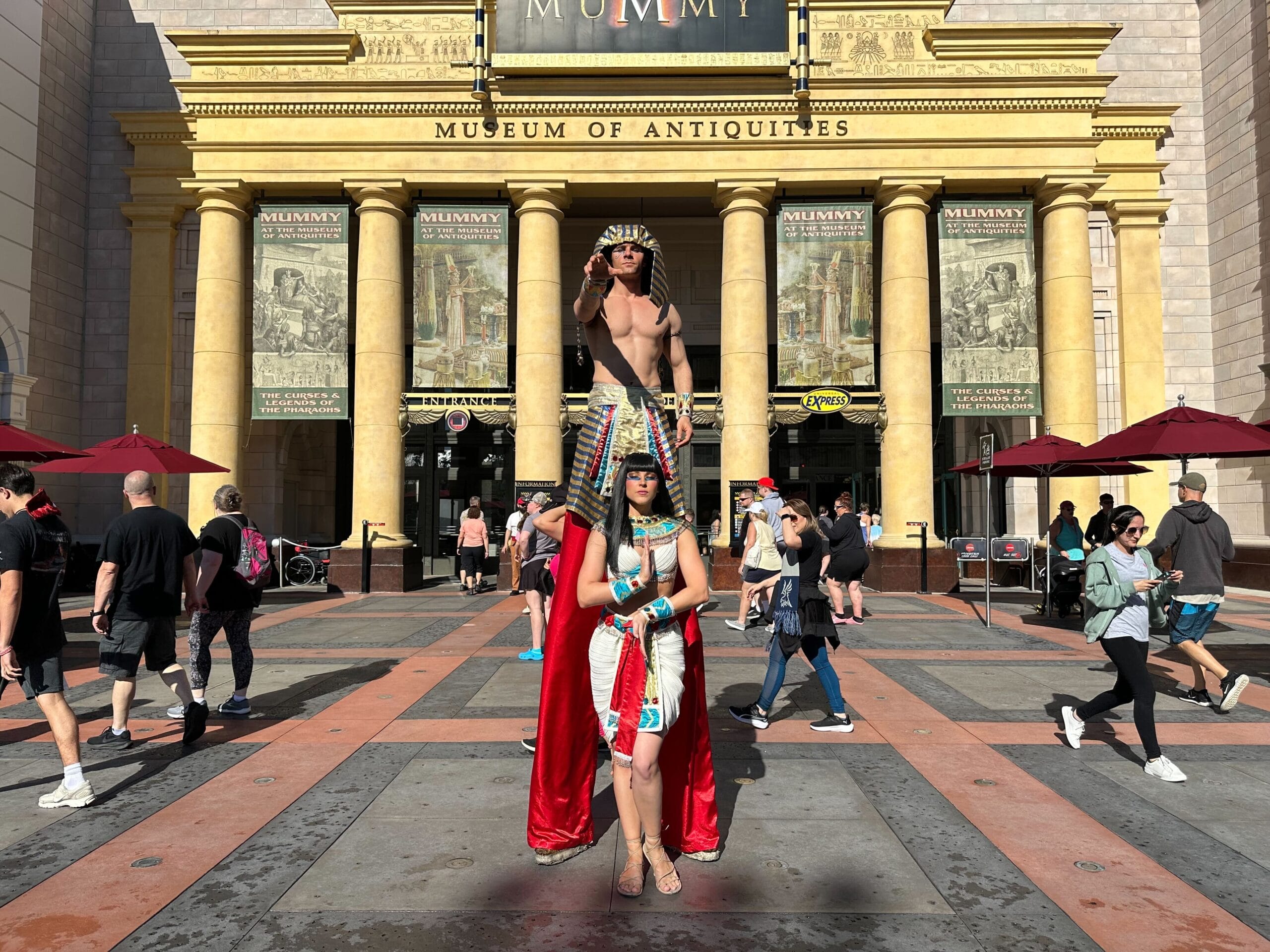Two people in Egyptian costumes pose by a museum, like actors from Revenge of the Mummy Ride, as crowds stroll in sunlight.