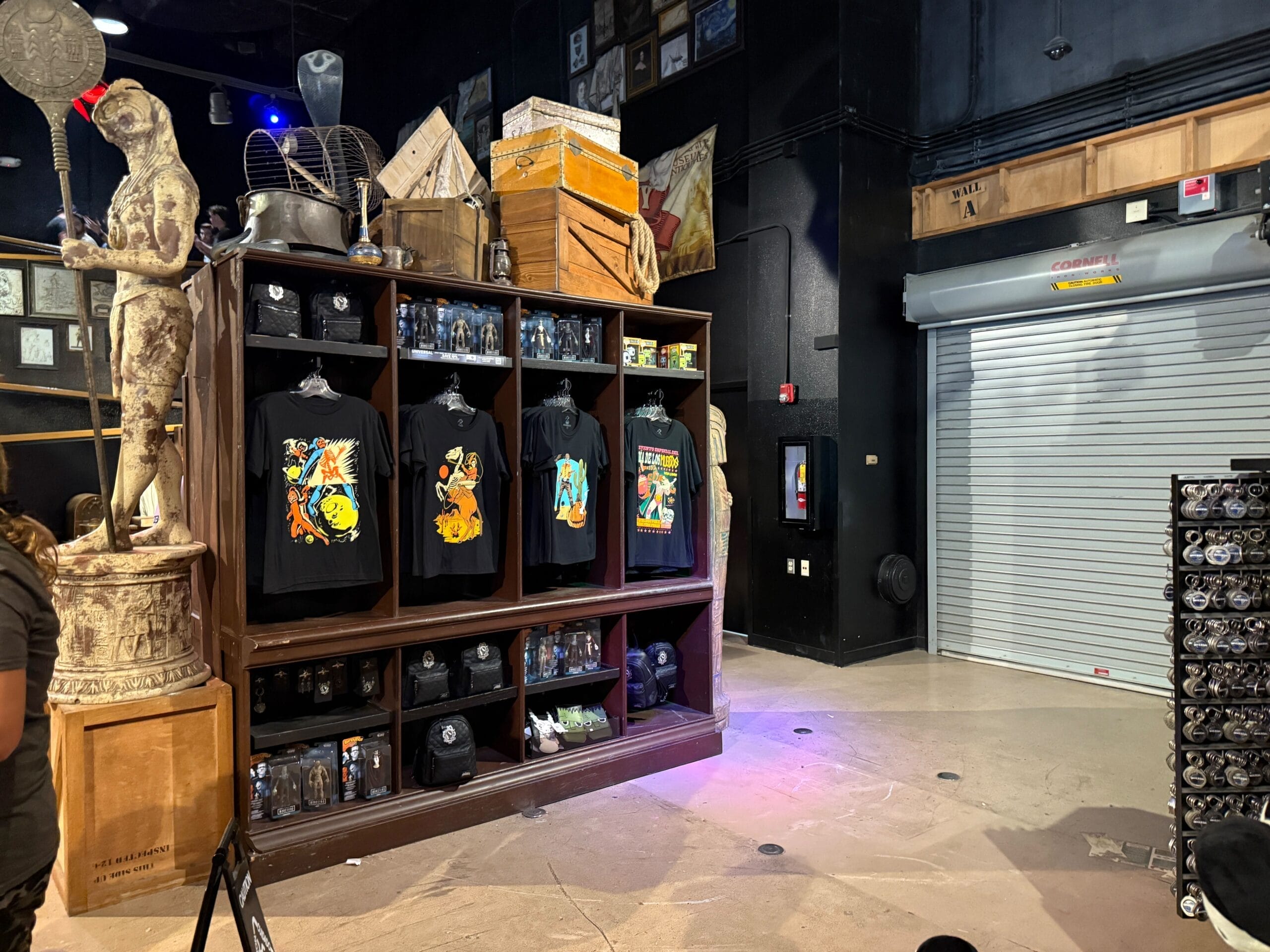 A Tribute Store display with graphic T-shirts, mugs, statues, and crates; a closed metal door is on the right.
