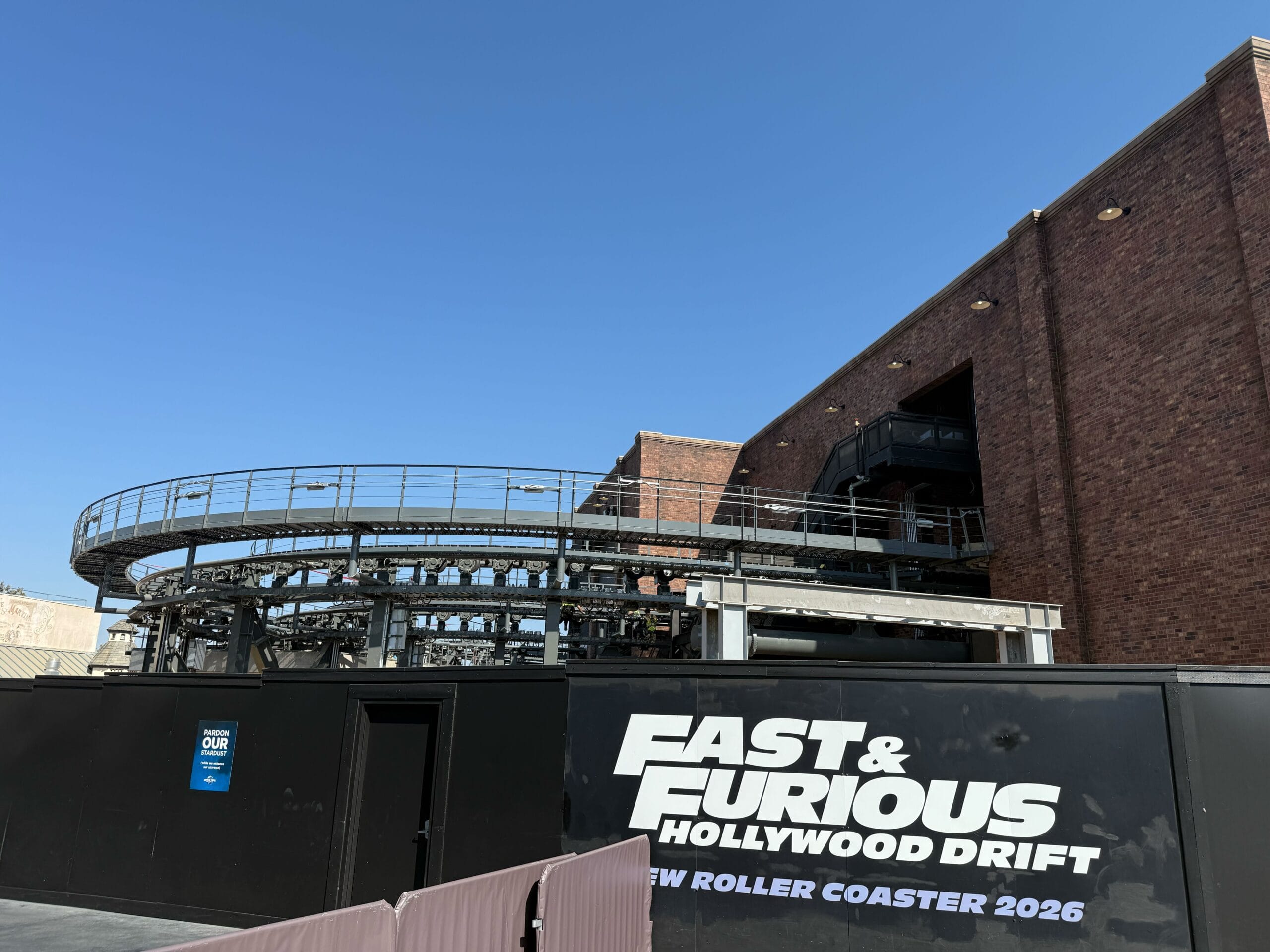 Construction site for Fast & Furious Hollywood Drift roller coaster with track and large brick building under a clear blue sky.