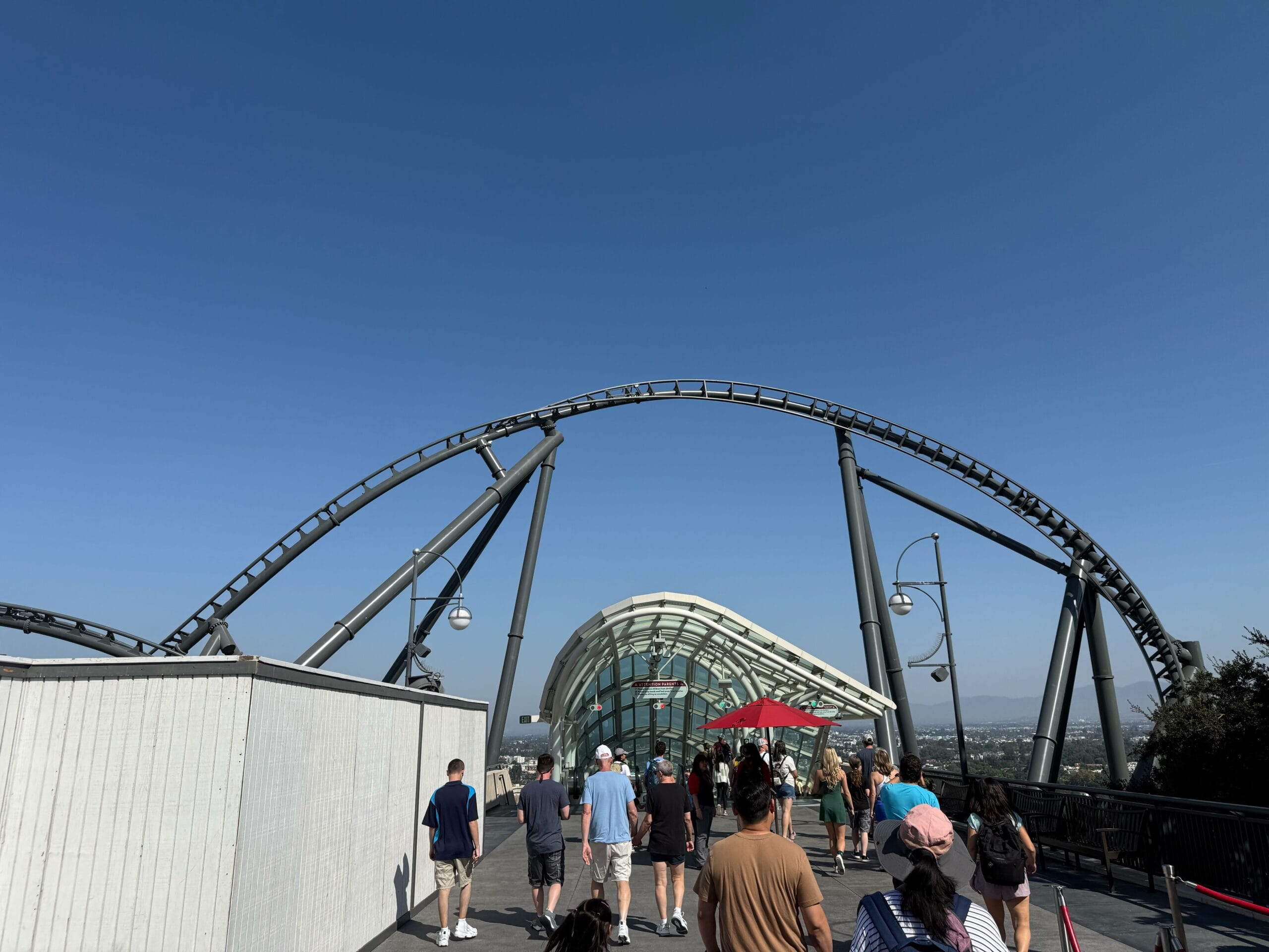 People walk under a roller coaster track next to a glass structure on a sunny day with a clear blue sky.