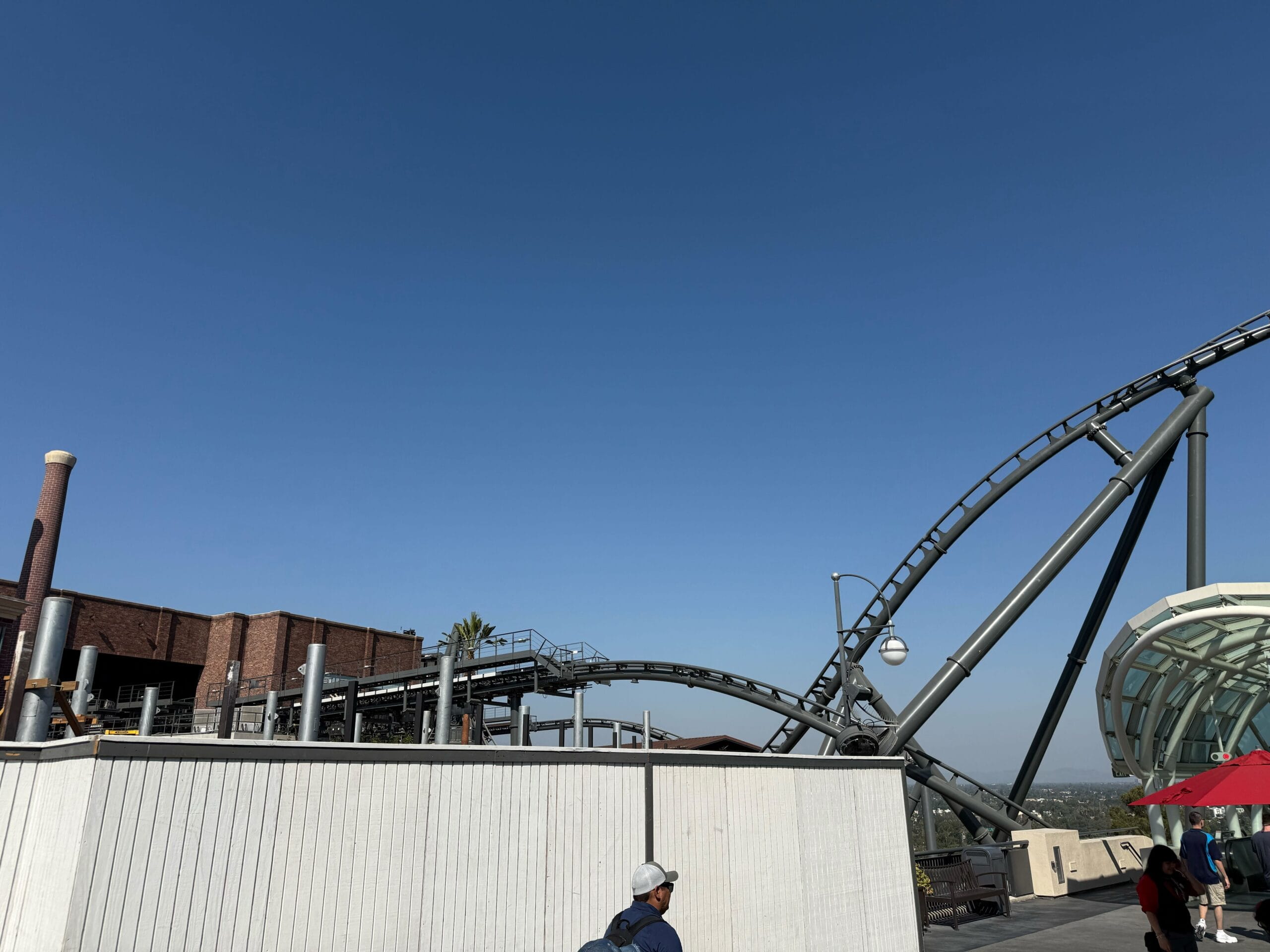 A roller coaster track curves above a white fence under a clear blue sky; a person in a hat walks nearby.