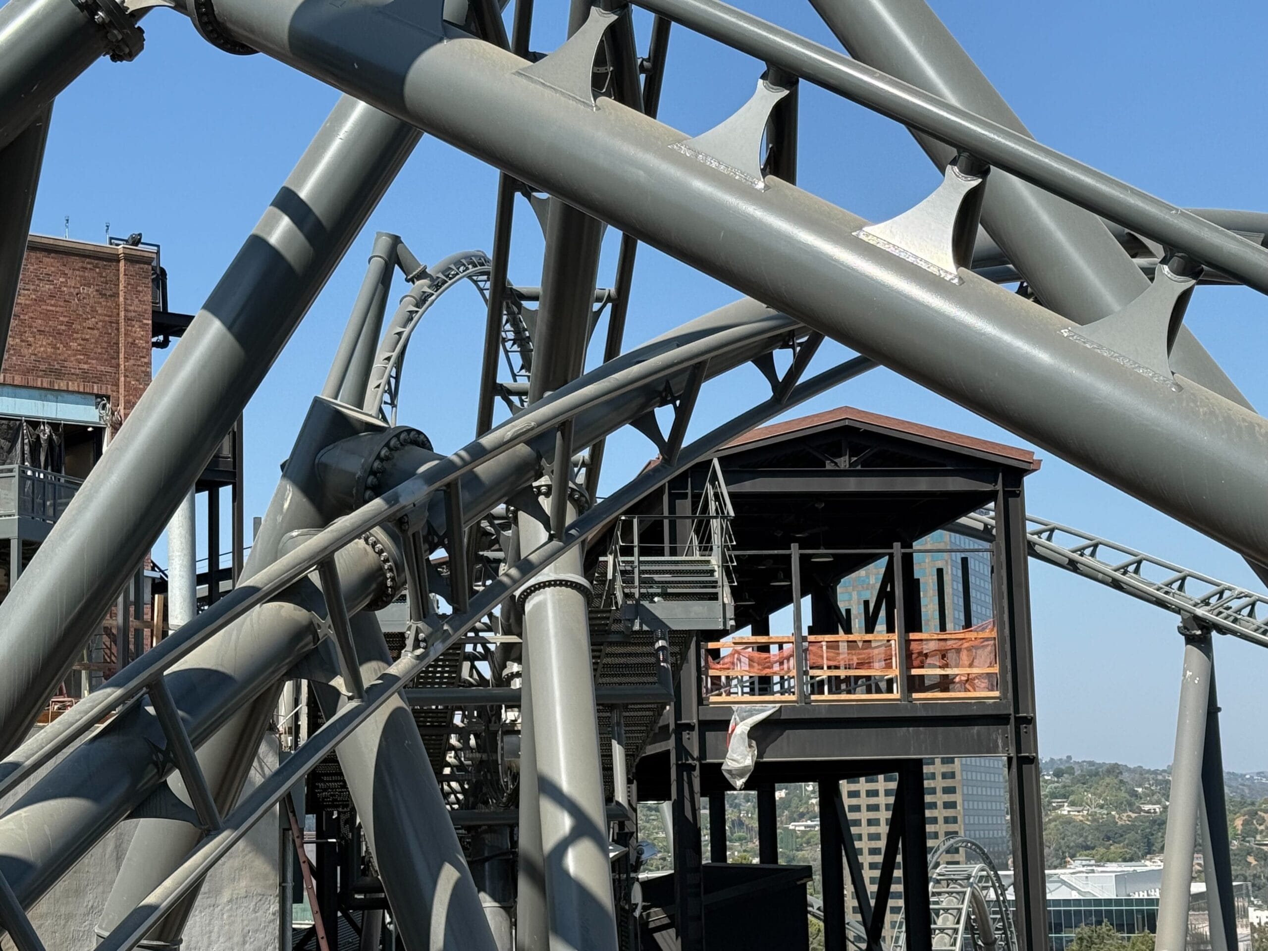 Gray roller coaster track curves near a station at Universal Studios Hollywood, cityscape and blue sky in the background.