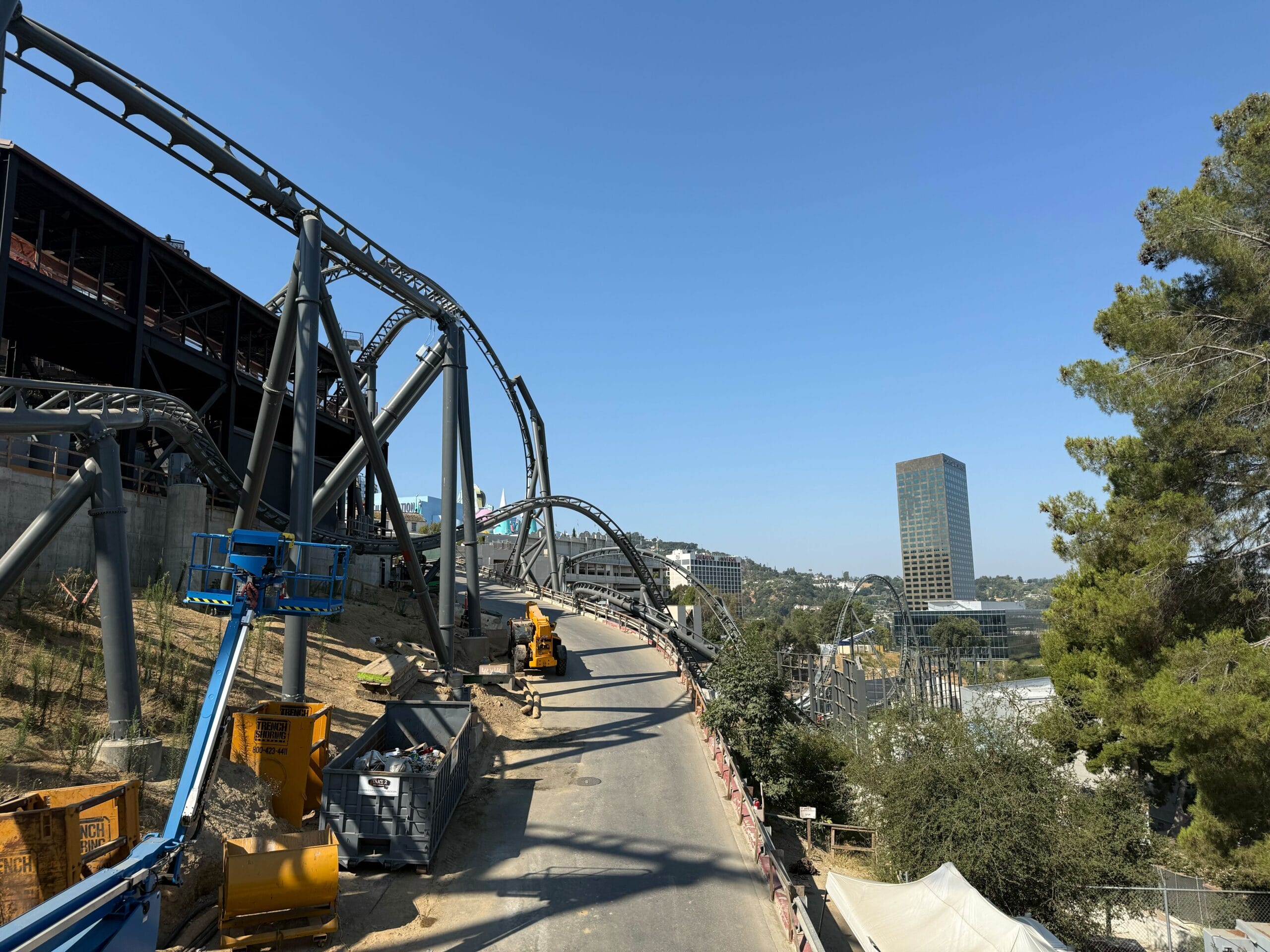 Roller coaster under construction at Universal Studios Hollywood, cranes and equipment visible, city buildings and trees behind.