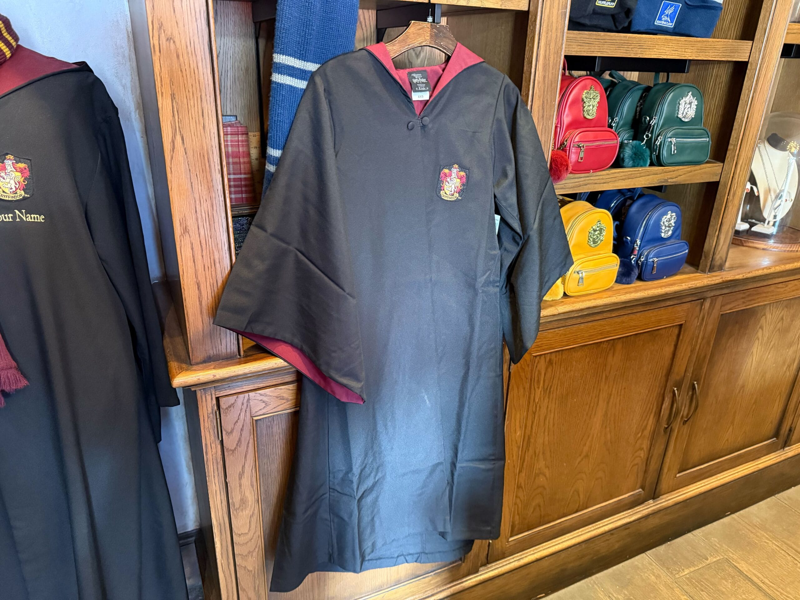 A Gryffindor youth robe hangs on display near shelves with colorful Harry Potter bags and scarves.