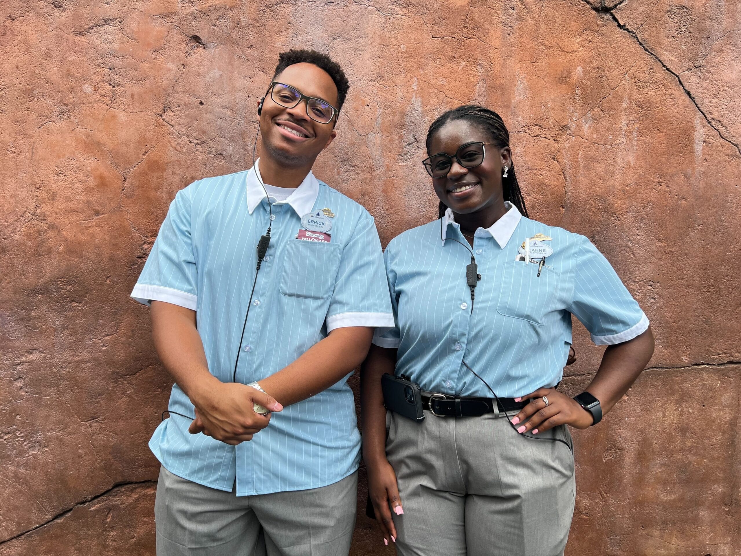 Two theme park Cast Member Coordinator Costumes employees smile in blue uniforms before a textured brown wall.