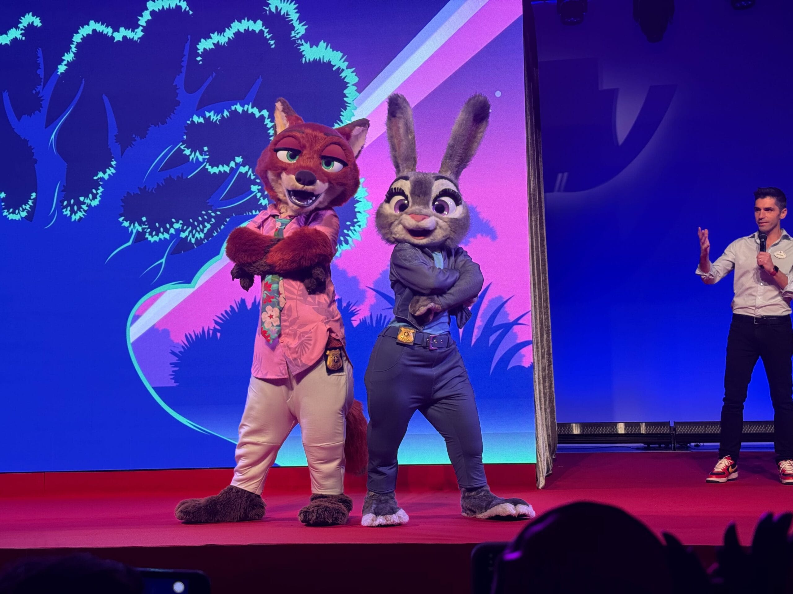 Nick Wilde and Judy Hopps mascots pose on stage at a Carrotvision Zootopia event, back-to-back with a presenter nearby.