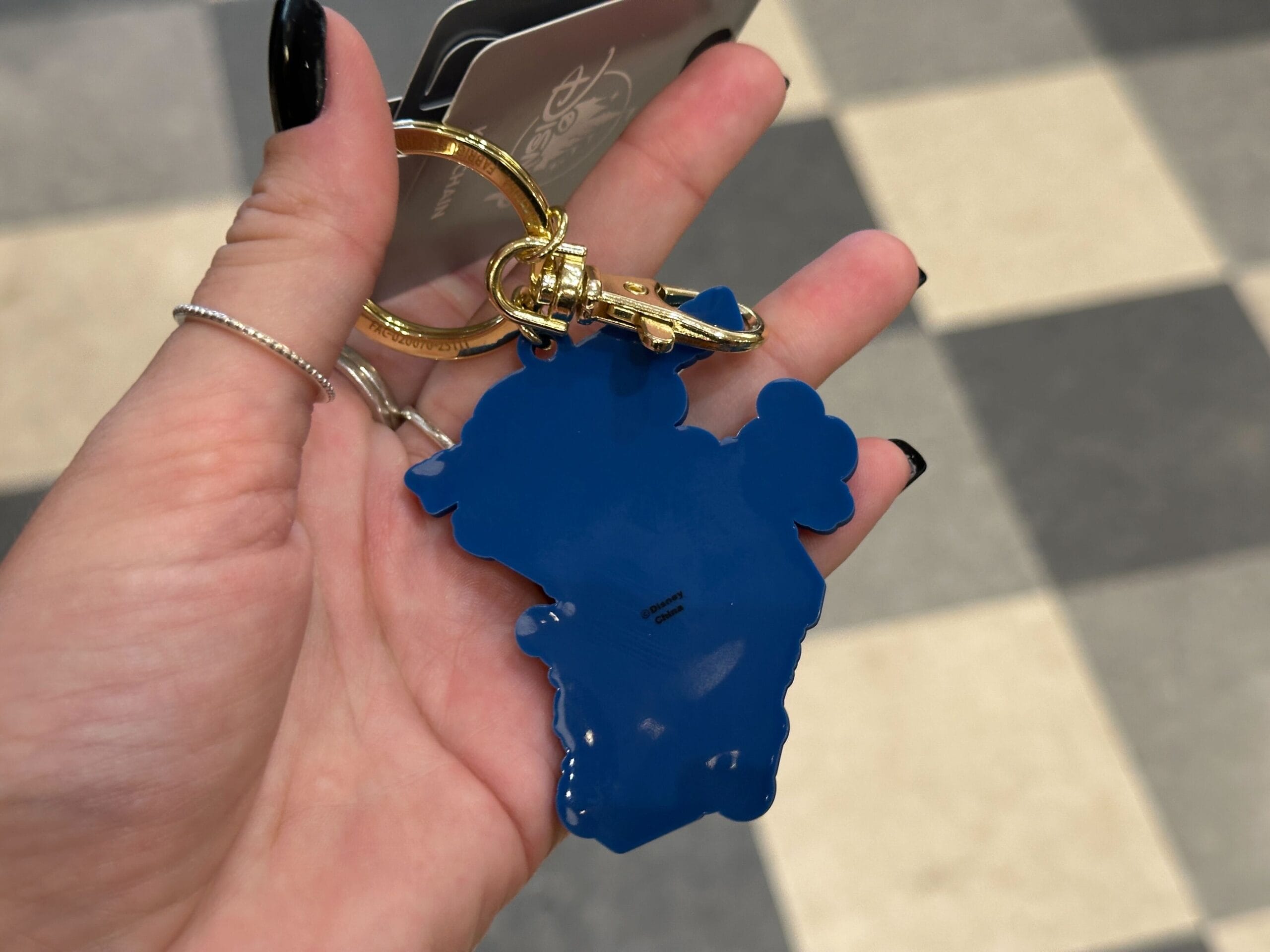 A hand holds a blue character-shaped keychain with a gold keyring over a checkered floor.