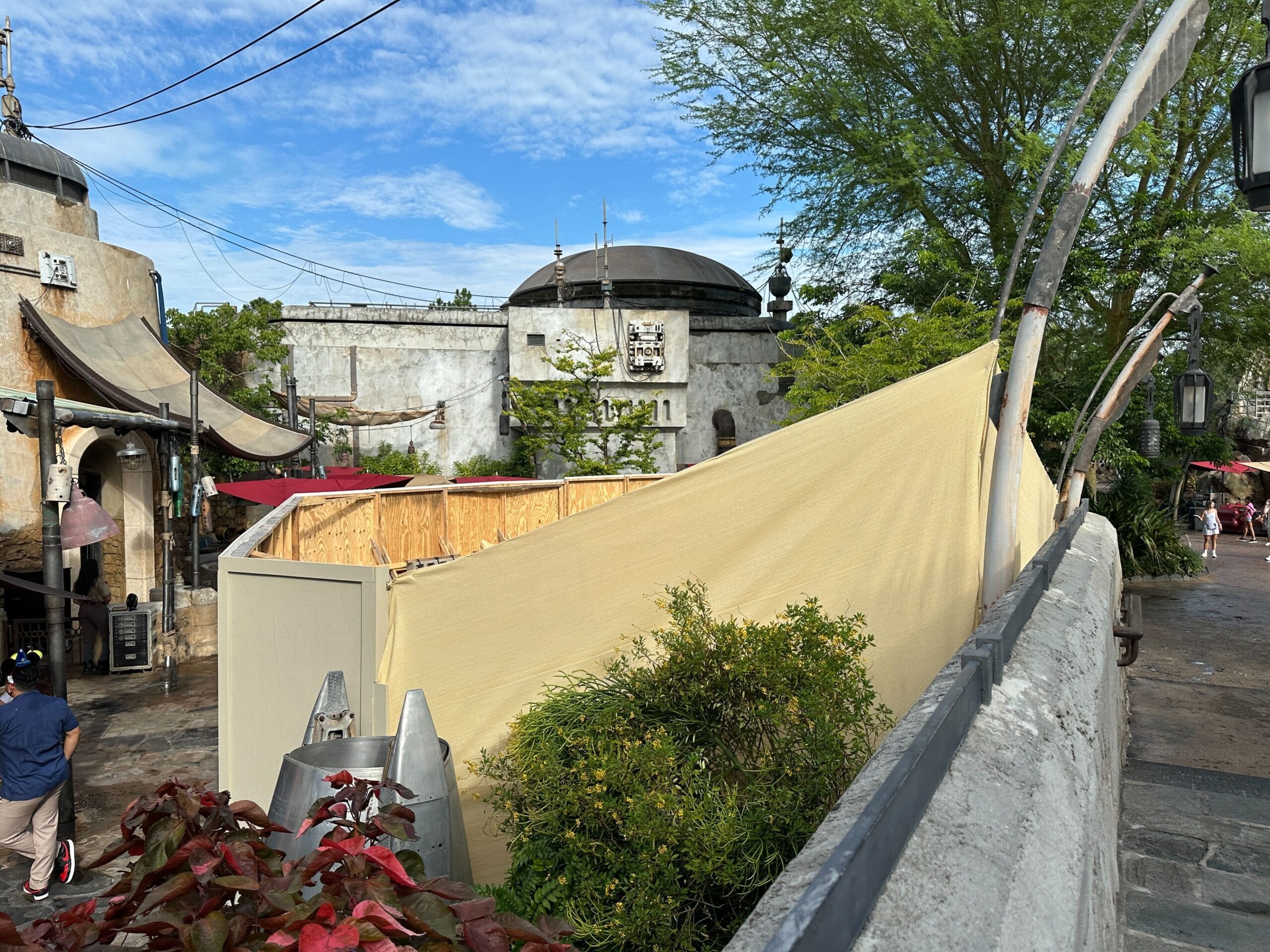 Construction walls with tan tarp and wood panels near stone buildings at Star Wars: Galaxy’s Edge surrounded by greenery.