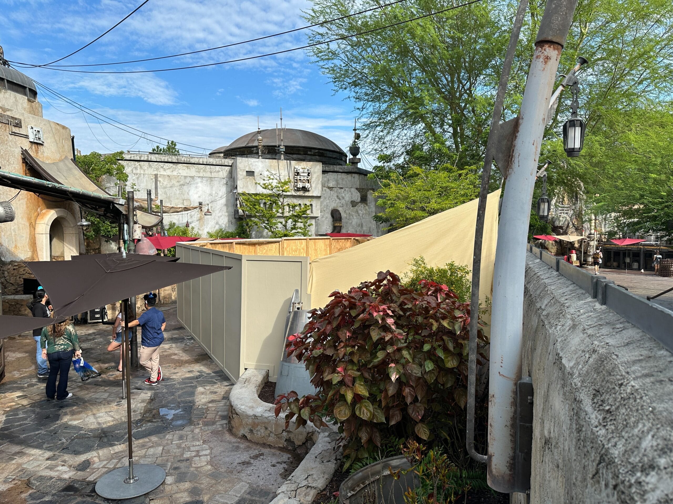 Outdoor scene with construction walls, plants, and people near Savi's Workshop in Star Wars: Galaxy's Edge under a blue sky.