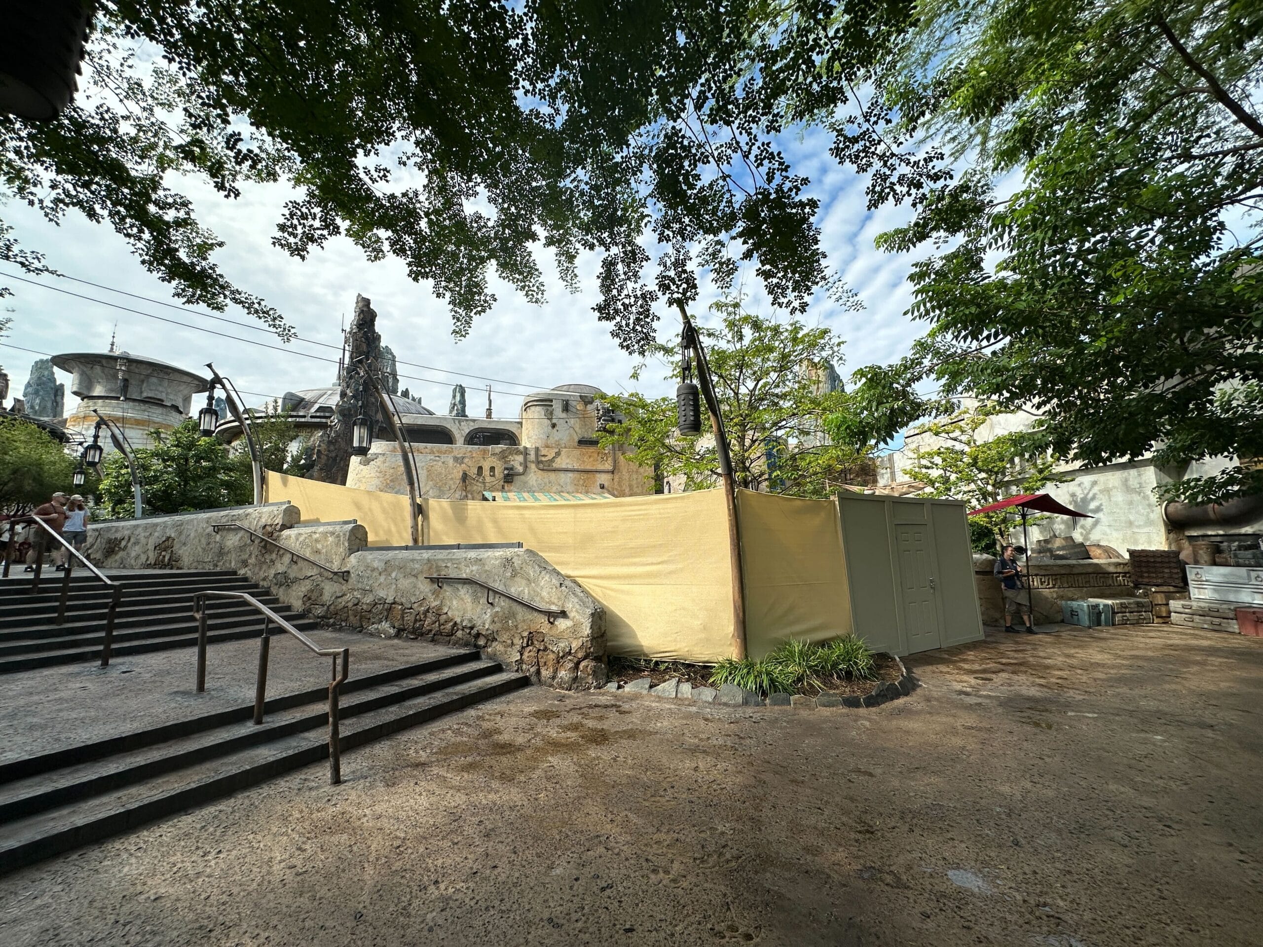 Yellow construction walls and a utility box block part of Star Wars: Galaxy's Edge, near trees and stone steps.