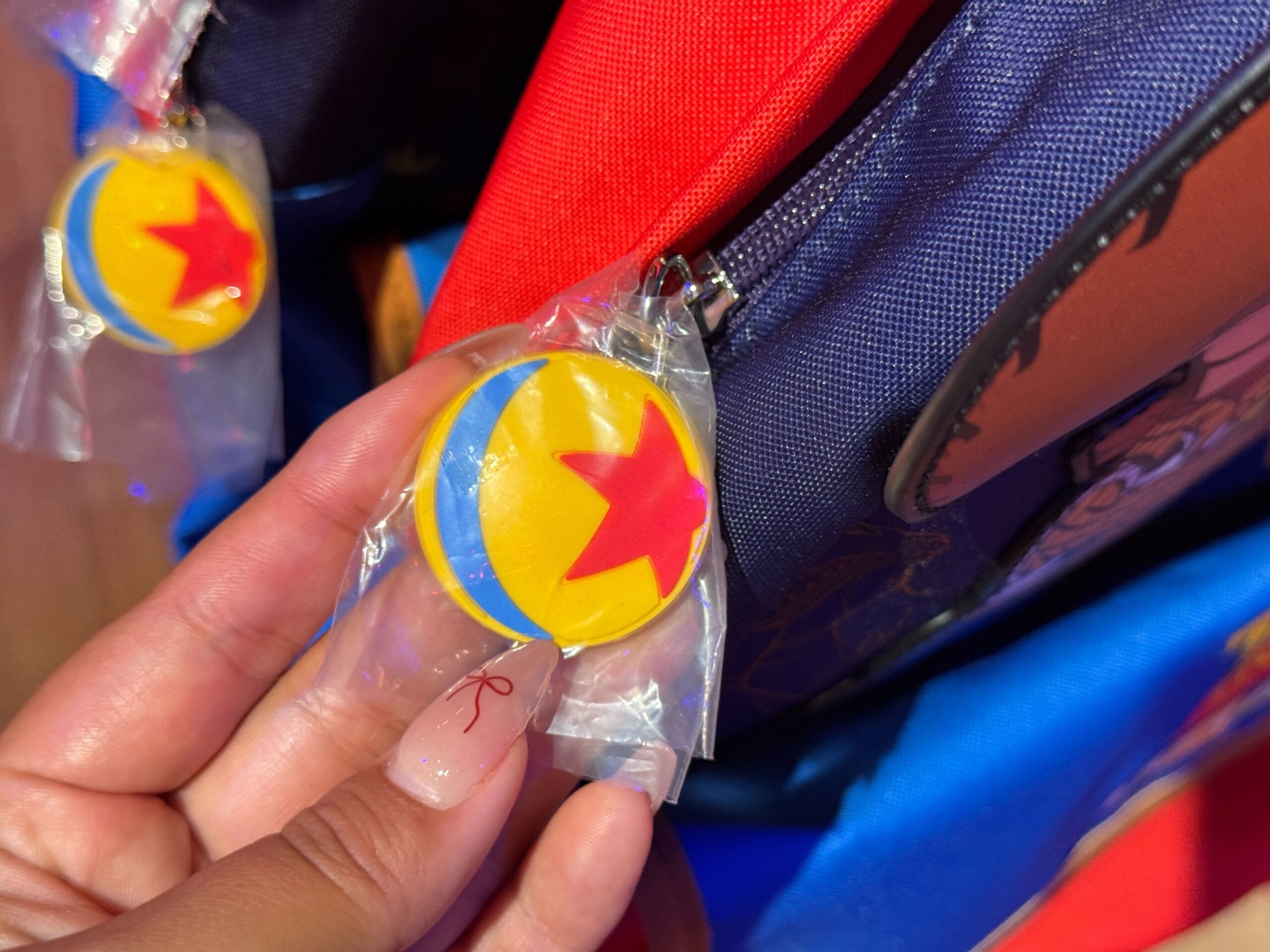 A hand holds a wrapped Pixar ball keychain near a blue and red backpack.
