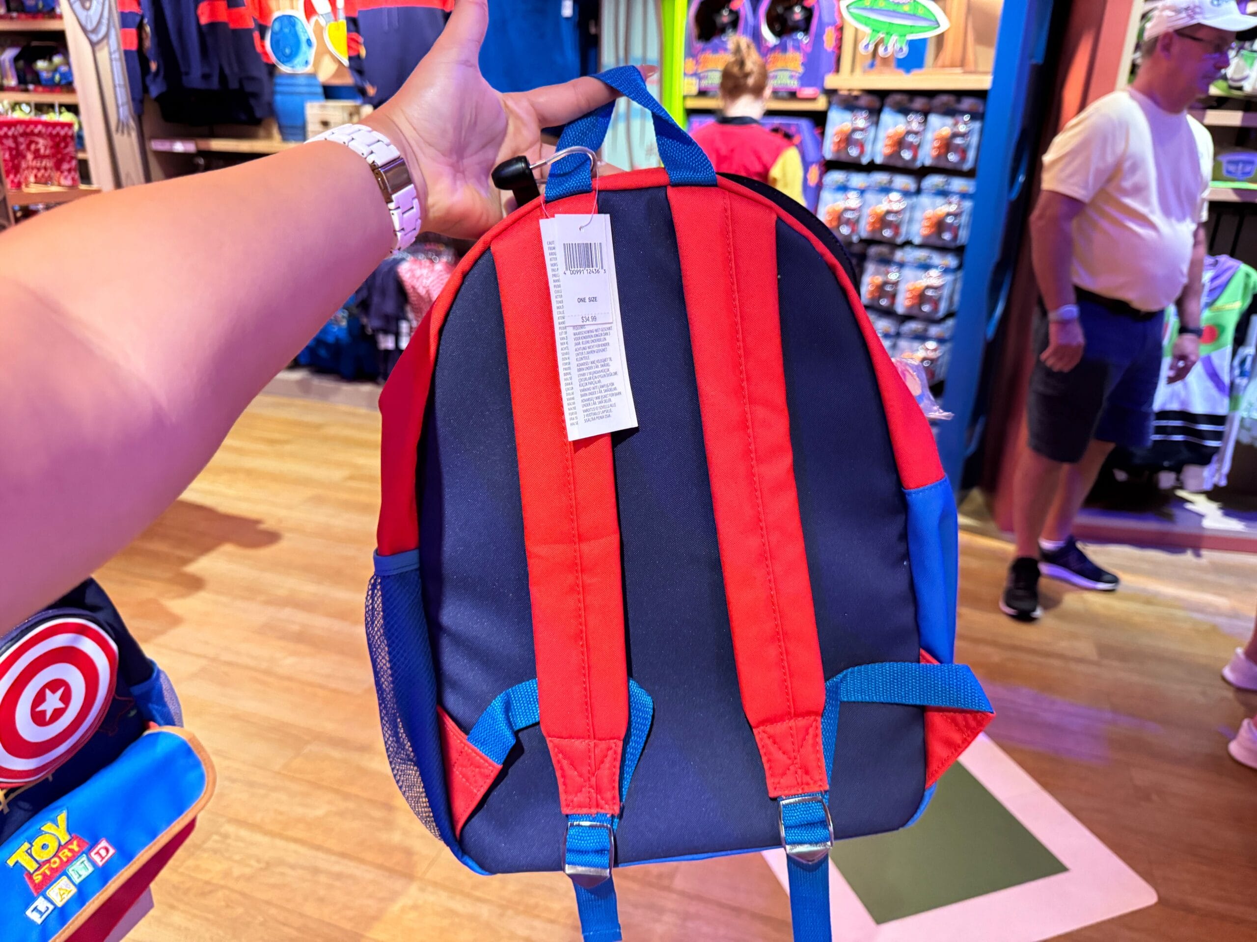 Person holding a blue and red backpack in a toy store with people and merchandise in the background.