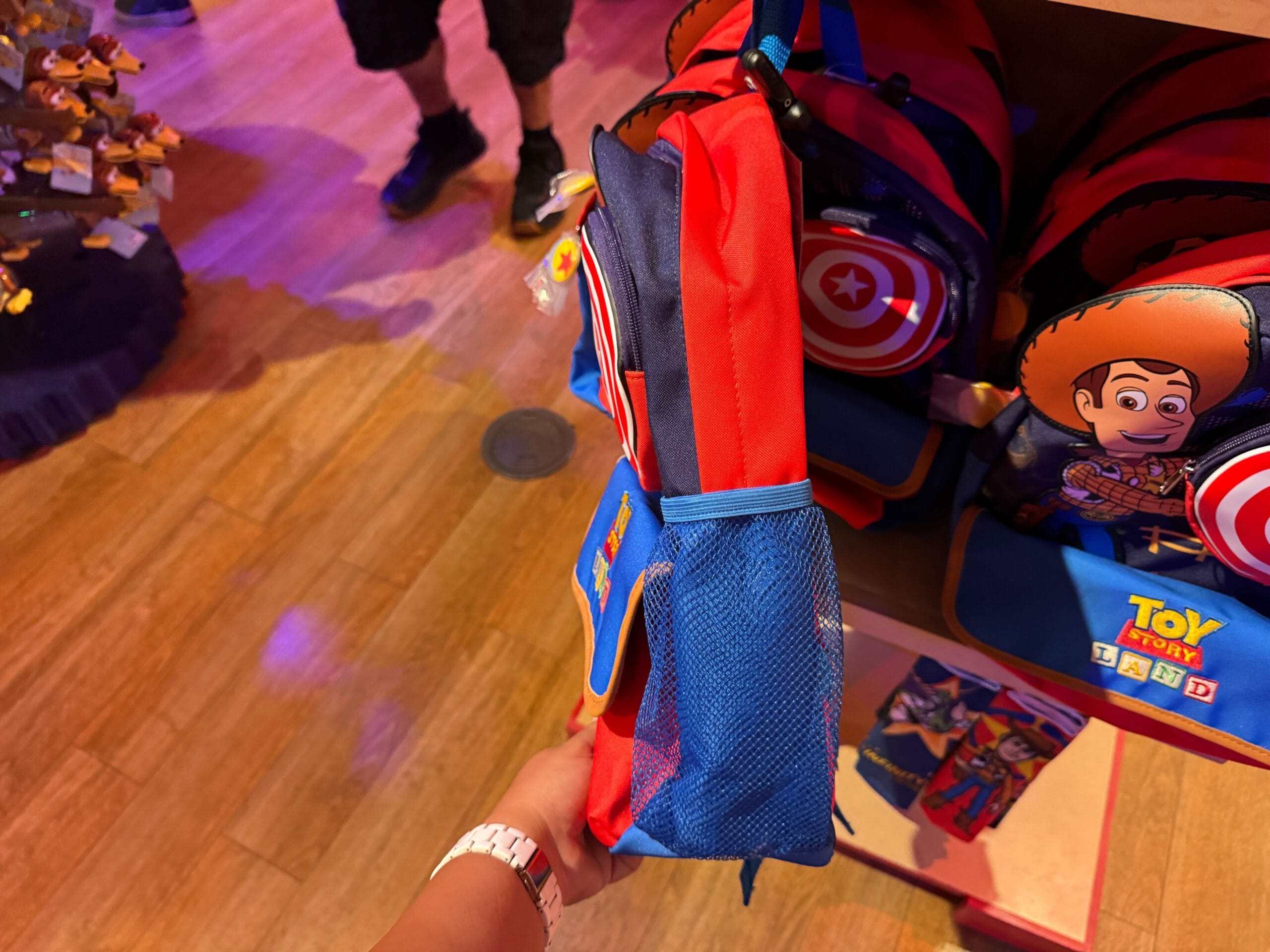 A hand holds a small Toy Story-themed backpack in a store with more backpacks in the background.