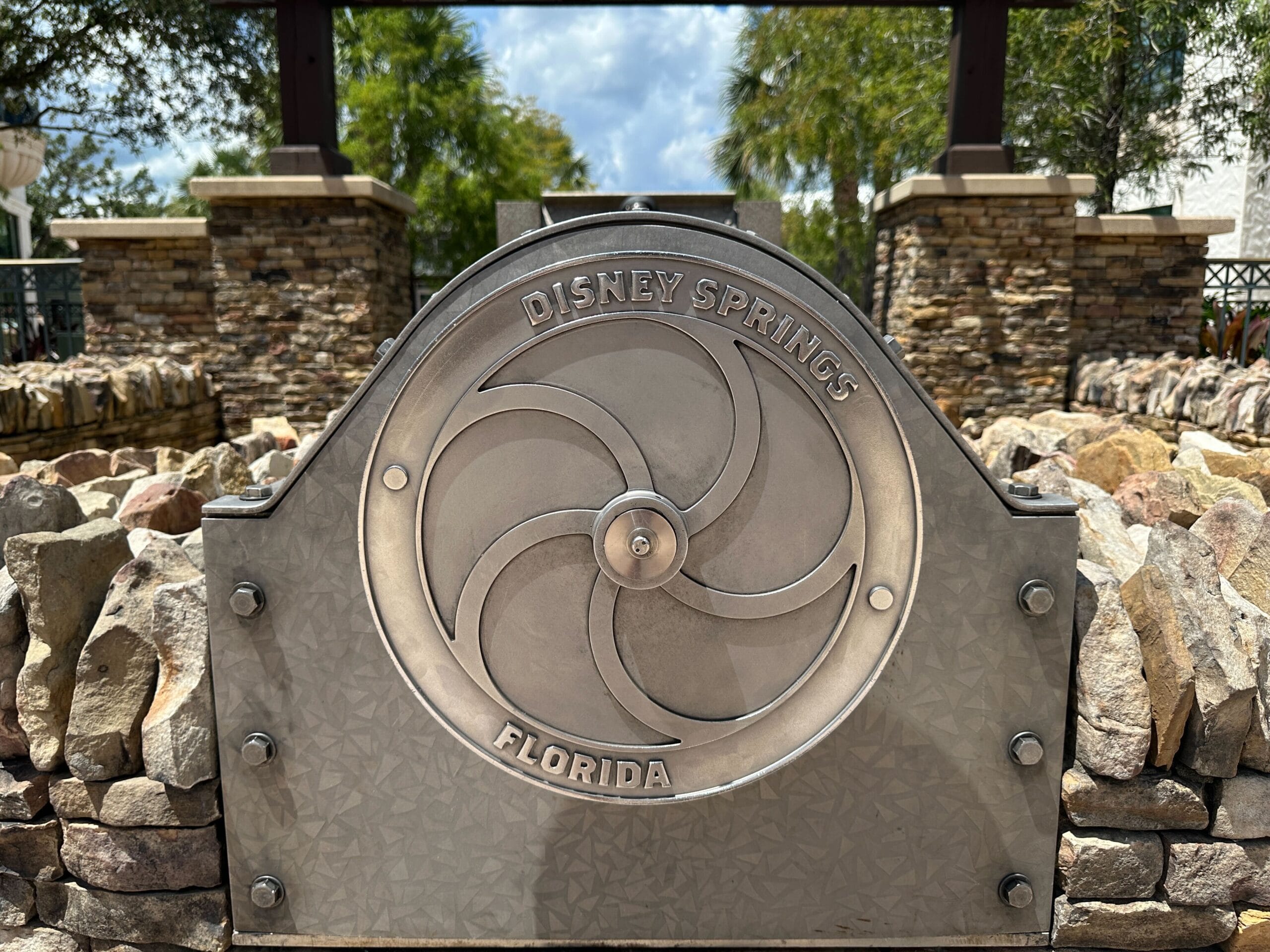 A metal sign reading "Disney Springs Florida" sits among rocks, trees, and an interactive water wheel on a sunny day.