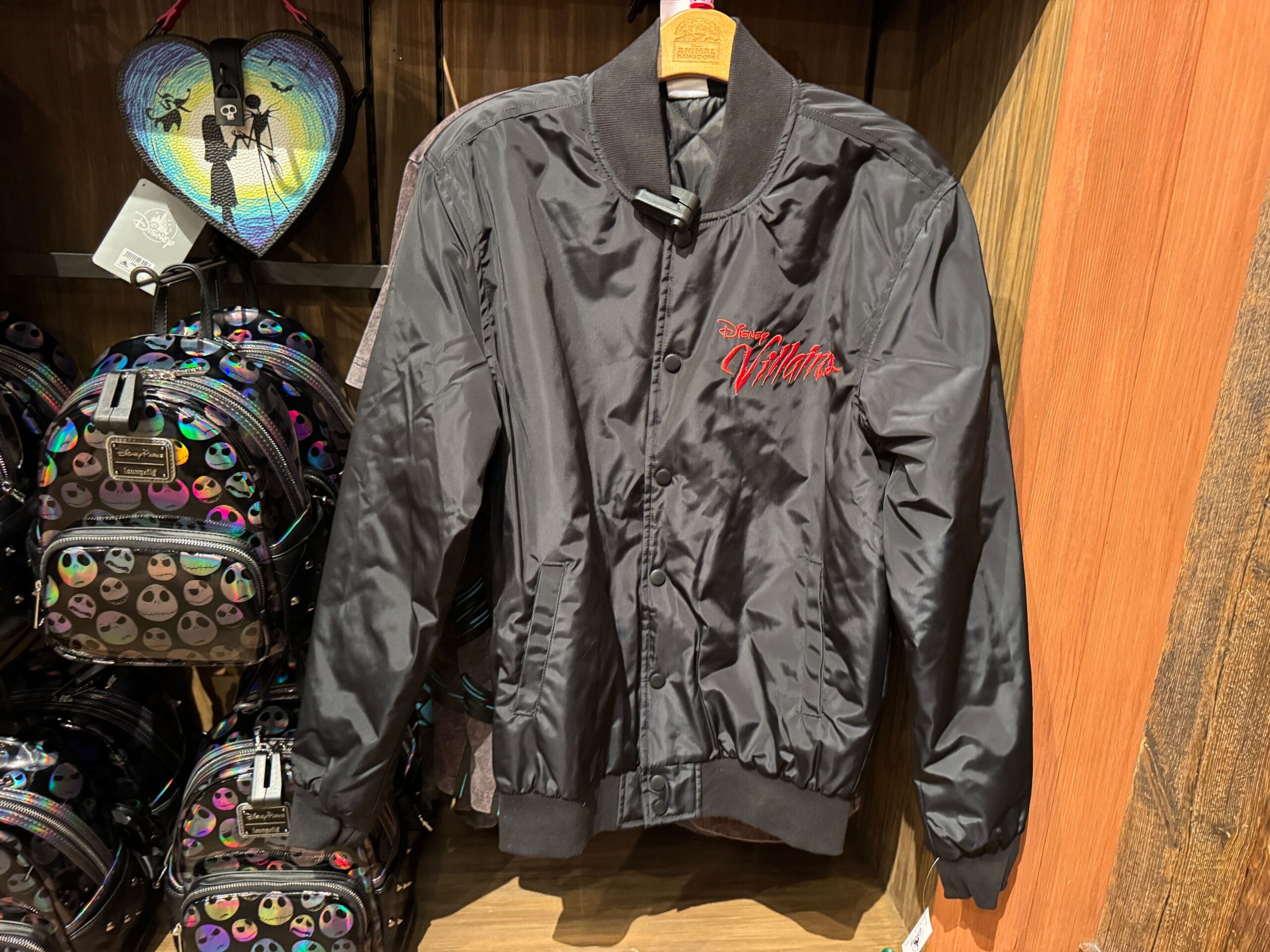 A black satin jacket with "Walt Disney Villains" embroidered in red on the chest, hanging on a wooden rack.