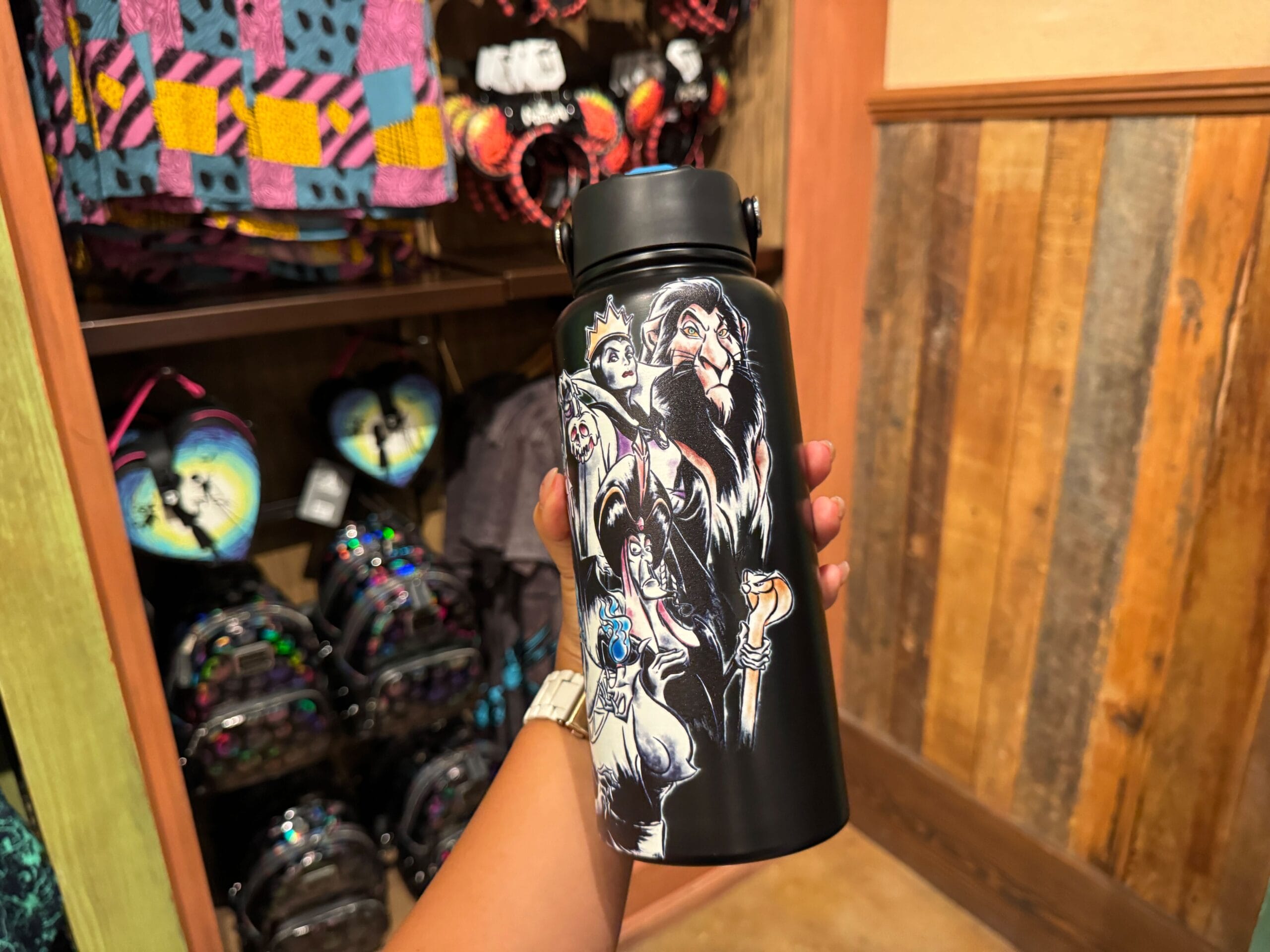 A hand holds Disney Villains merchandise water bottle in a store at Walt Disney World with colorful backpacks on shelves.