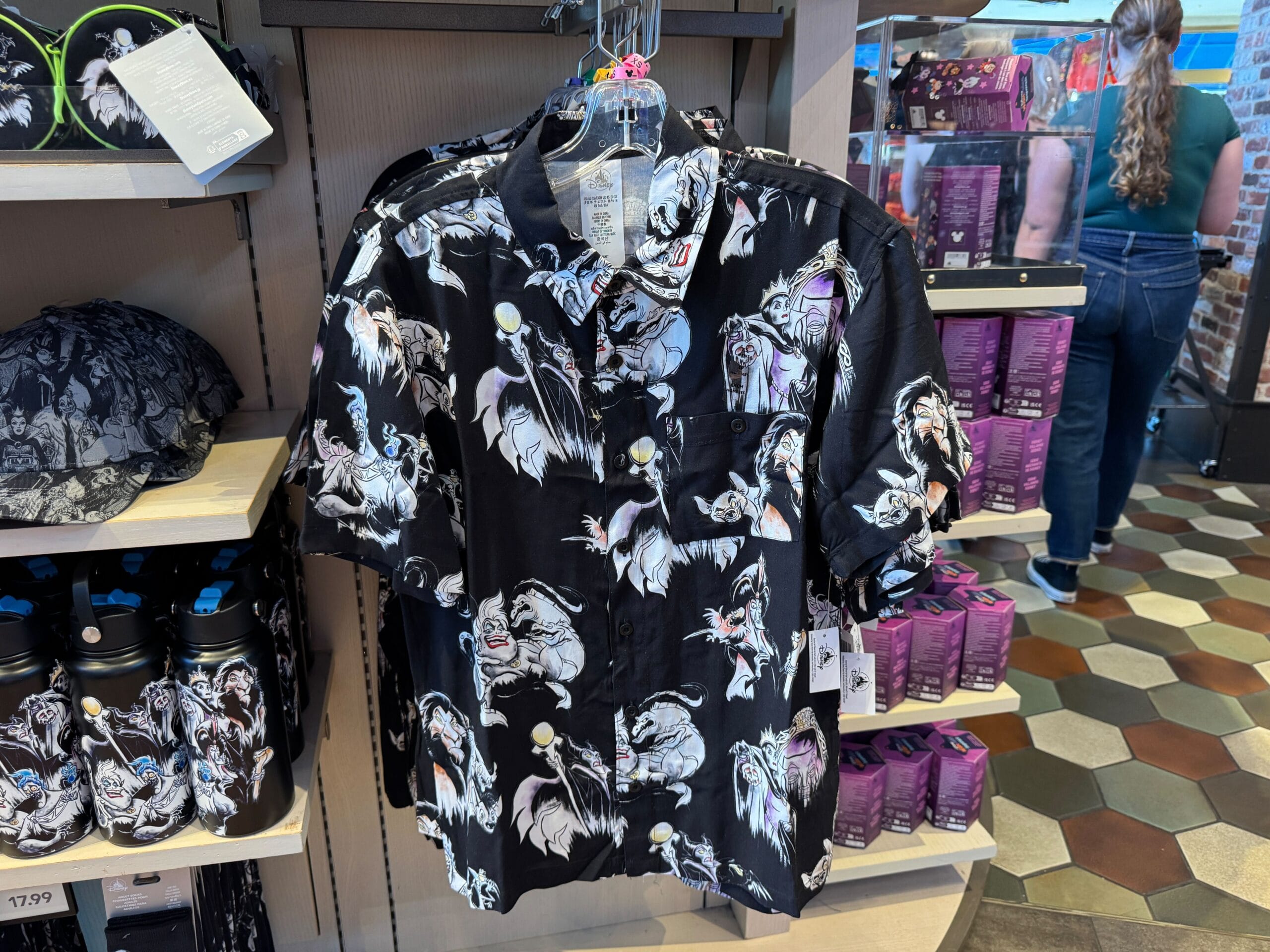 Black short-sleeve shirt with Disney villain prints displayed in a store, surrounded by hats and merchandise.