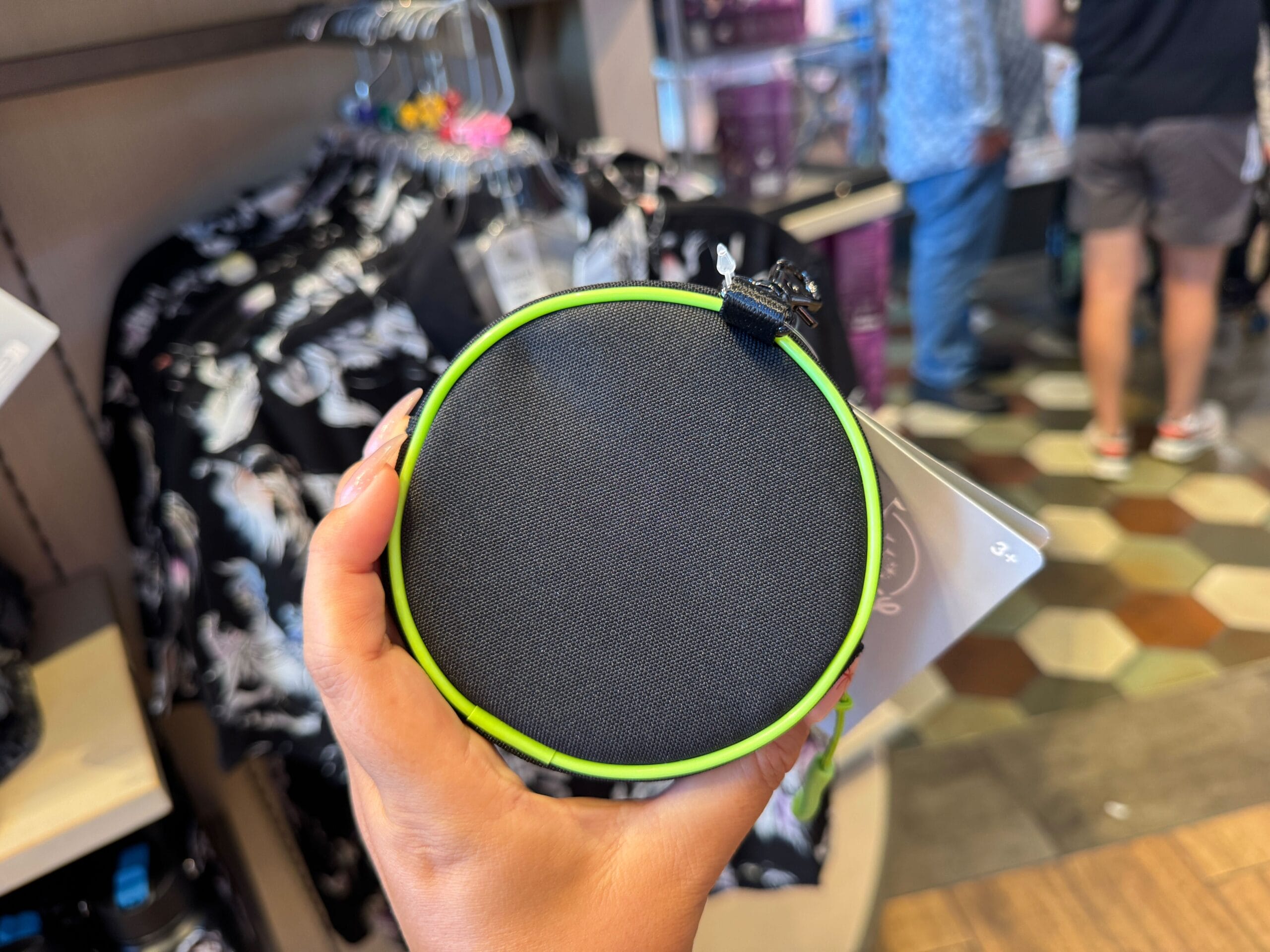 Hand holding a round, black zippered pouch with green edge—Disney Villains merchandise—inside a store with hexagon tiles.