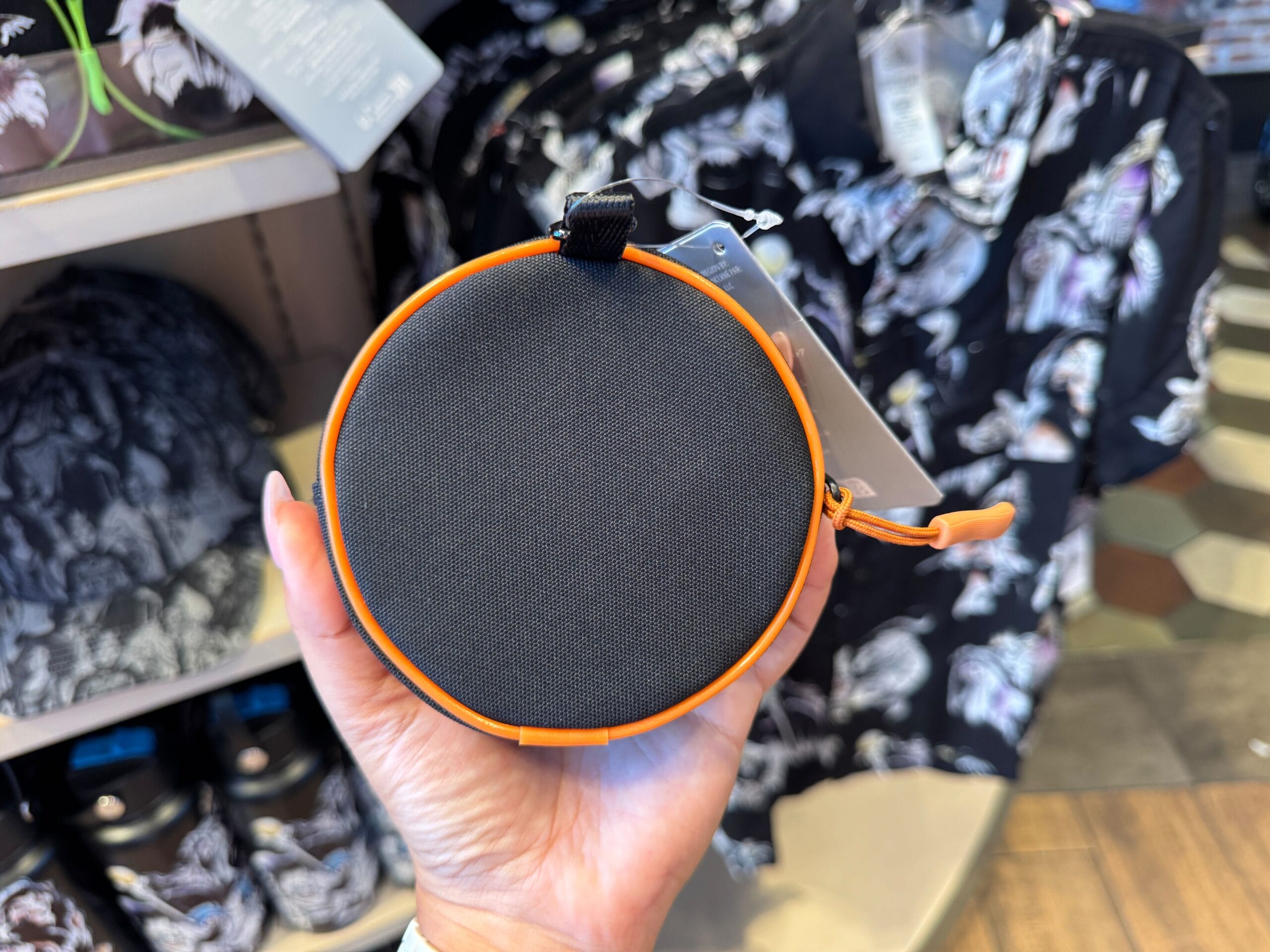 A hand holds a round, black and orange zippered pouch—Disney Villains Merchandise—amid patterned shirts in a retail store.
