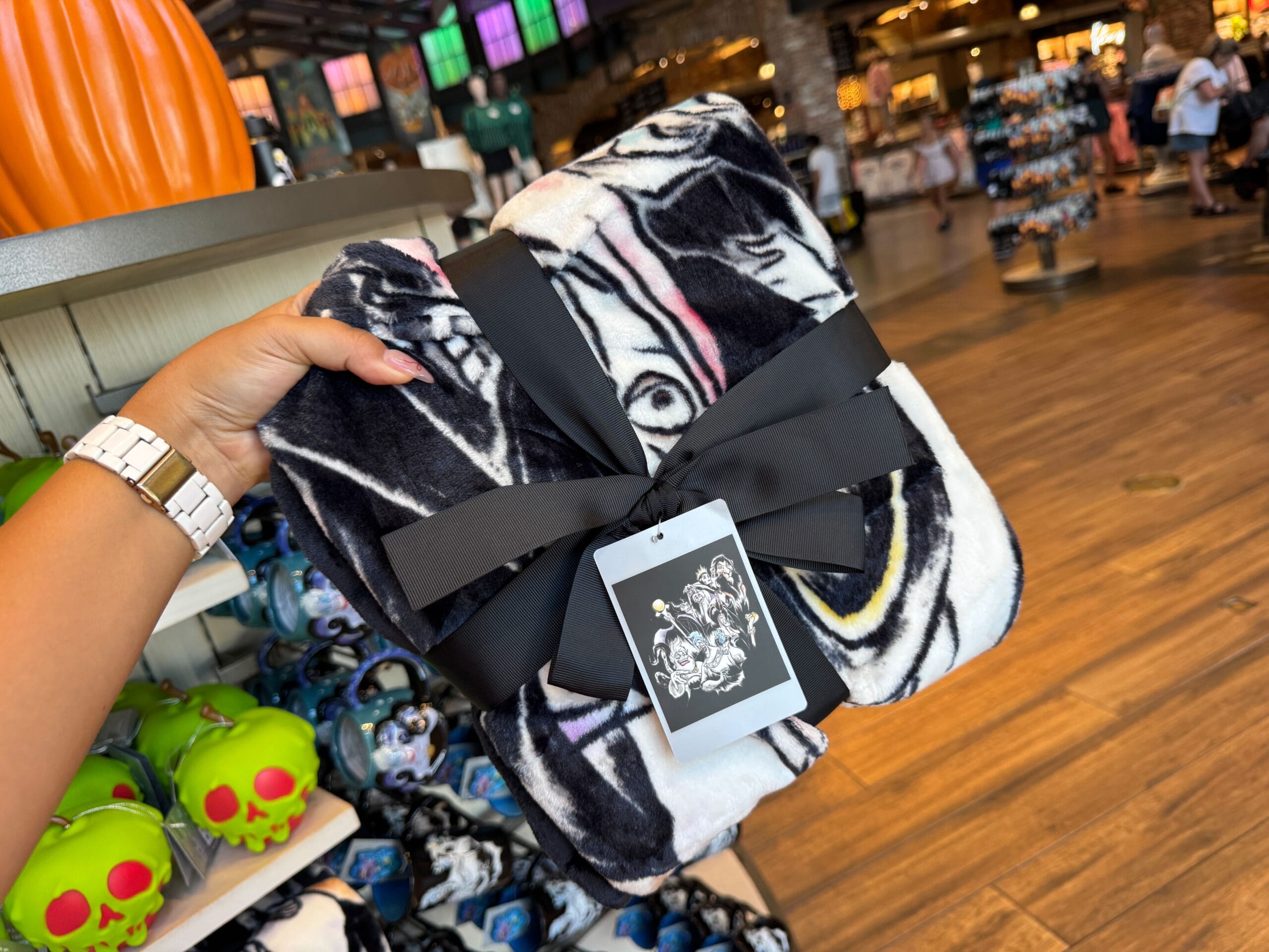 A hand holds a folded black and white blanket with a black ribbon, near Disney Villains merchandise in-store.
