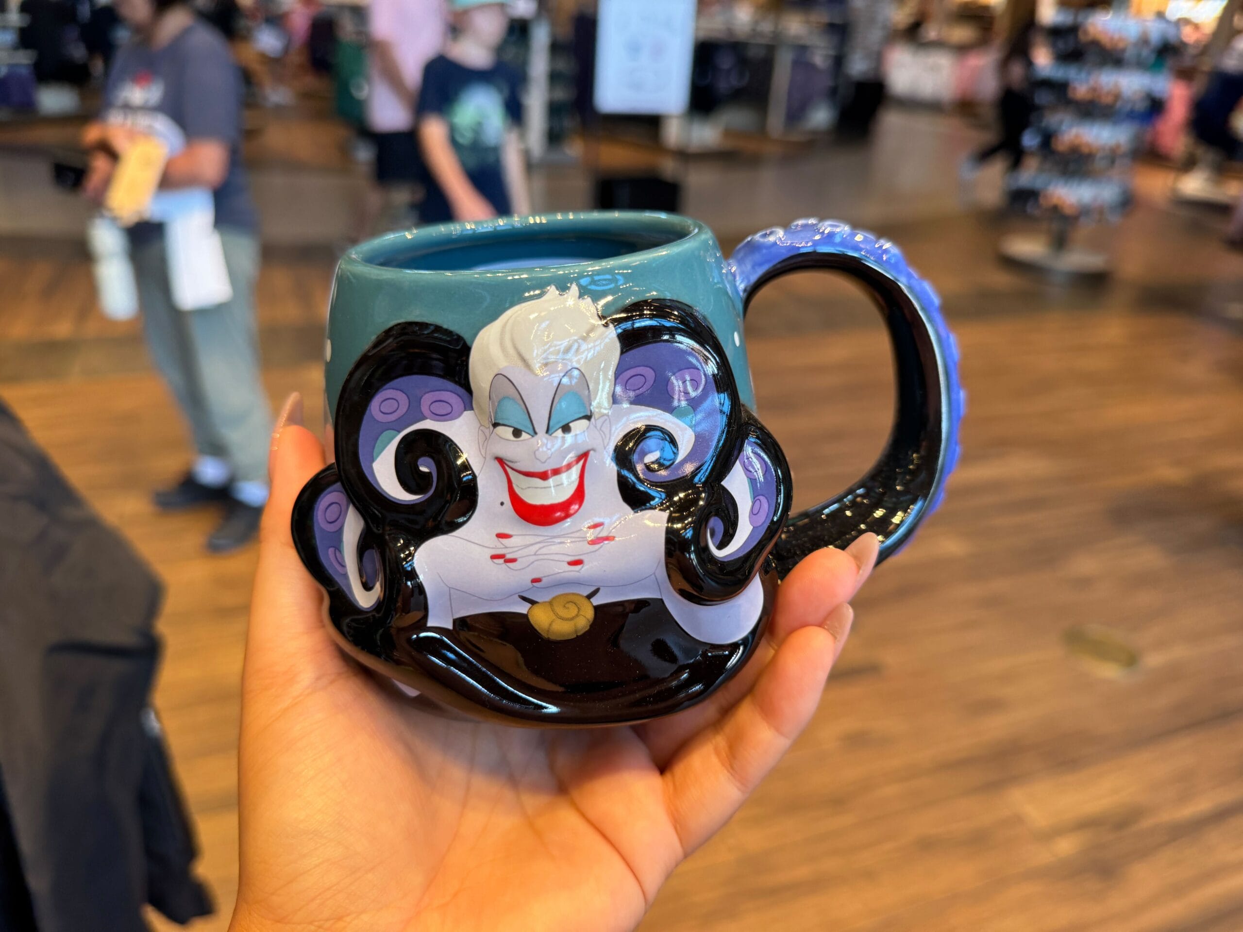 Hand holding a Disney Villains merchandise mug featuring Ursula at Walt Disney World, with a store and people in the background.