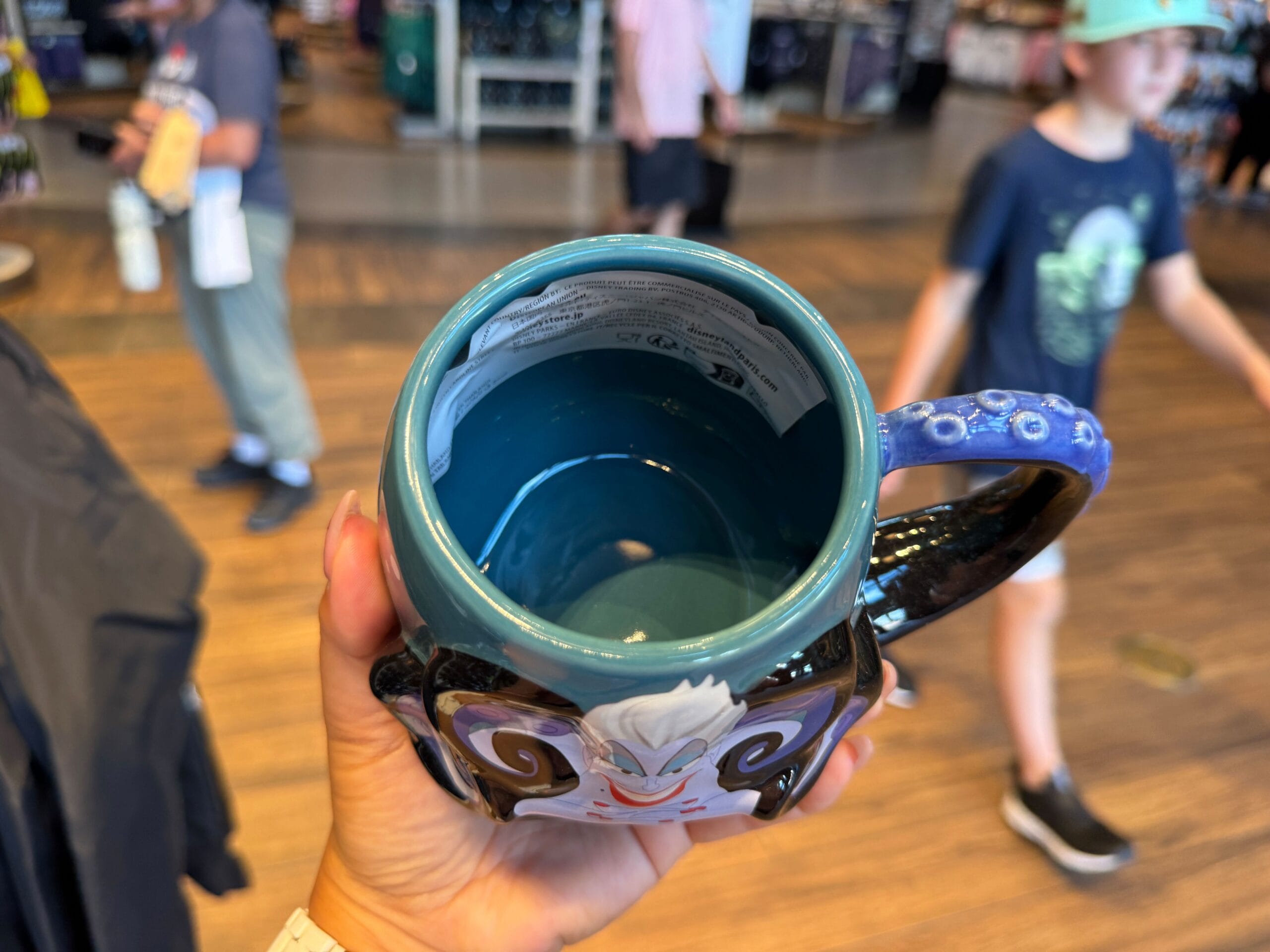 A hand holds a decorative blue mug; Disney Villains Merchandise lines the shelves as shoppers browse in the background.