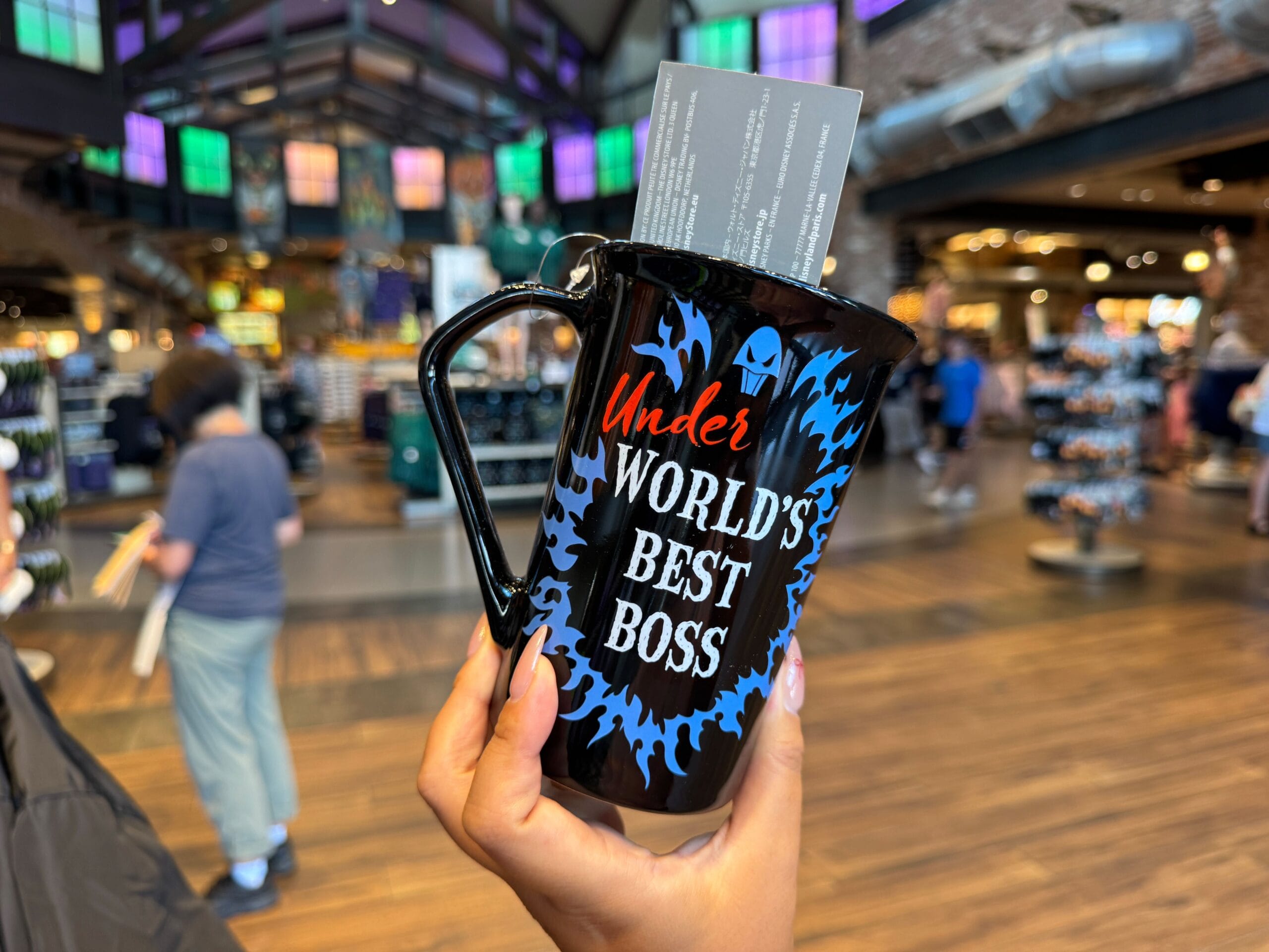 A hand holds a black mug with "Under WORLD'S BEST BOSS" in a colorful store filled with Disney Villains merchandise.