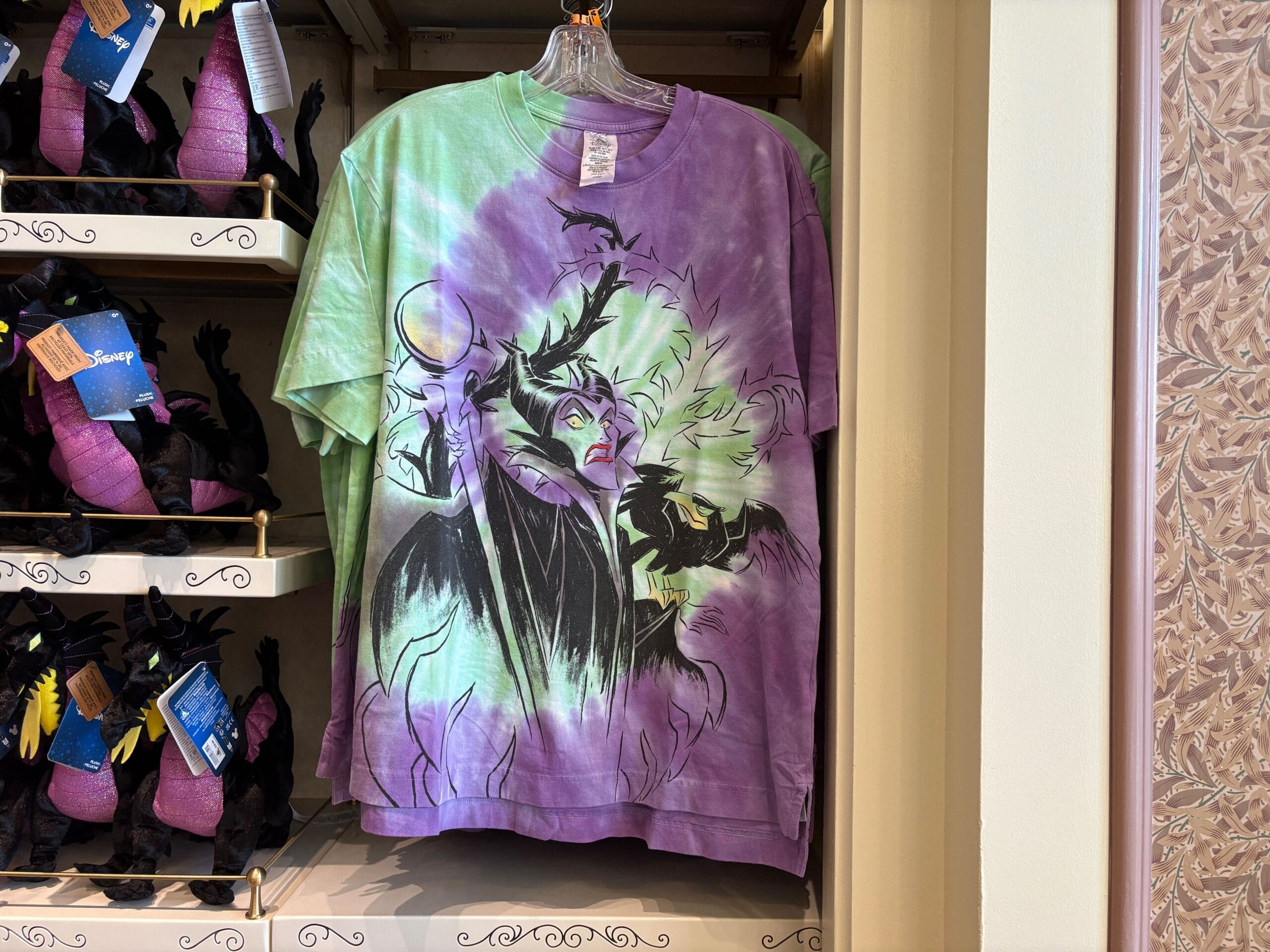 Tie-dye t-shirt with Maleficent and her raven, part of Disney Villains merchandise, displayed by plush dragon toys.