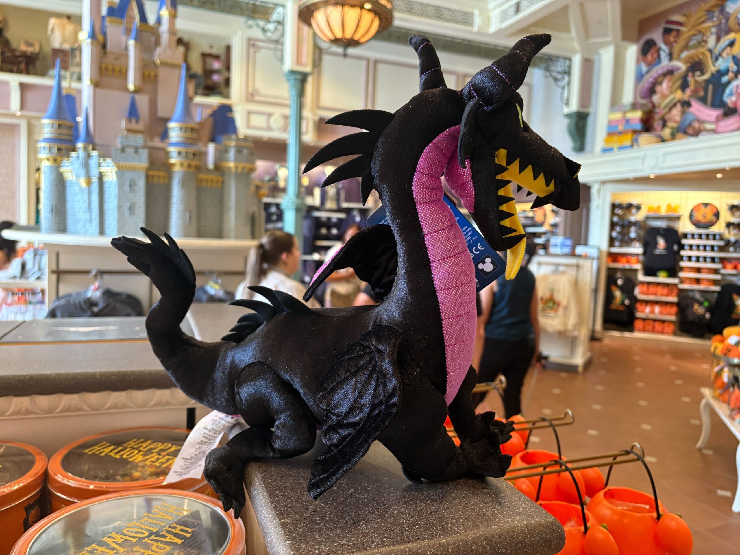 A plush black and purple dragon toy sits among Disney Villains Merchandise on a Halloween 2025 castle display shelf.