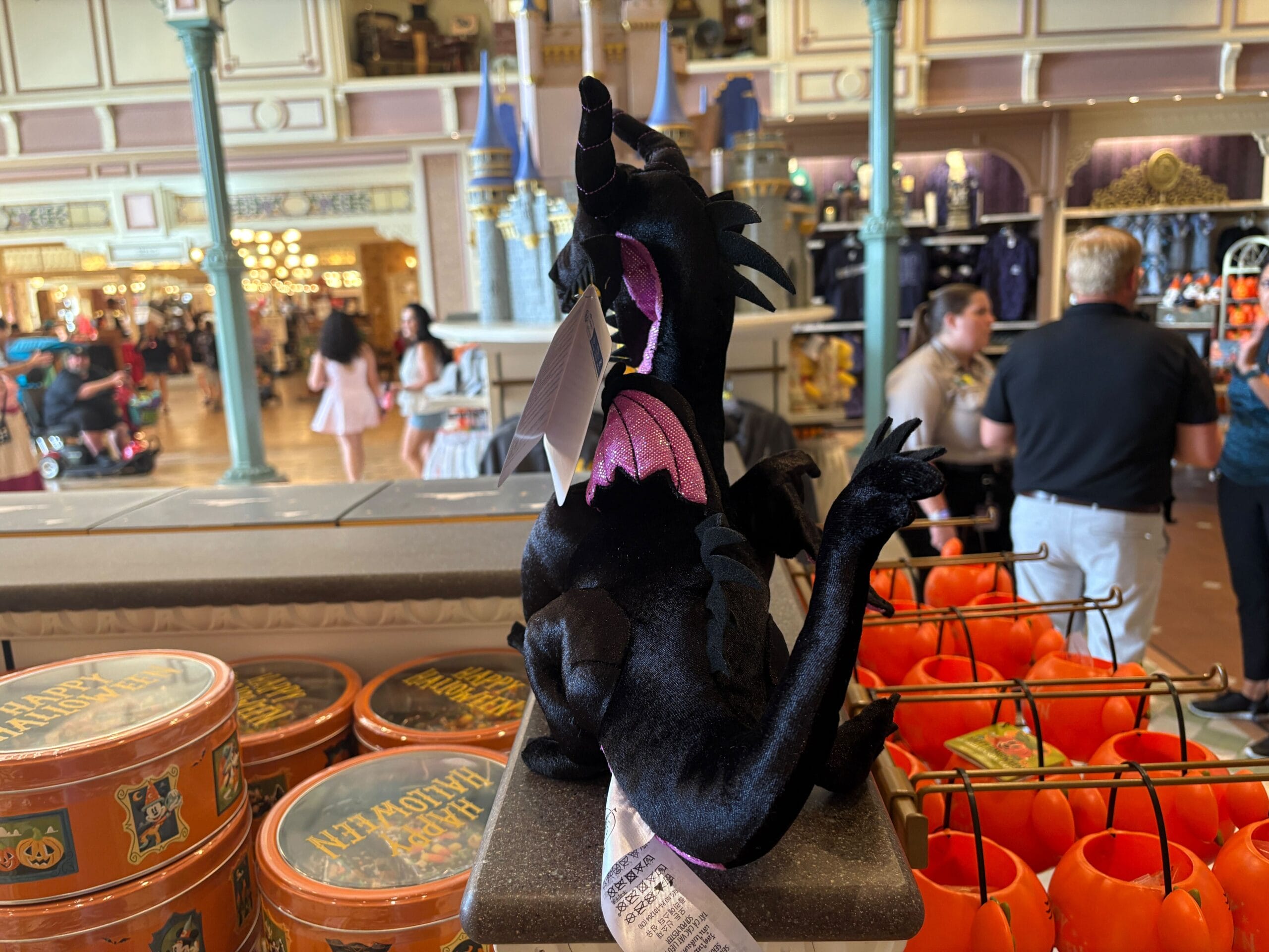 A black dragon plush, part of Disney Villains merchandise, sits on display amid Halloween decorations and shoppers.
