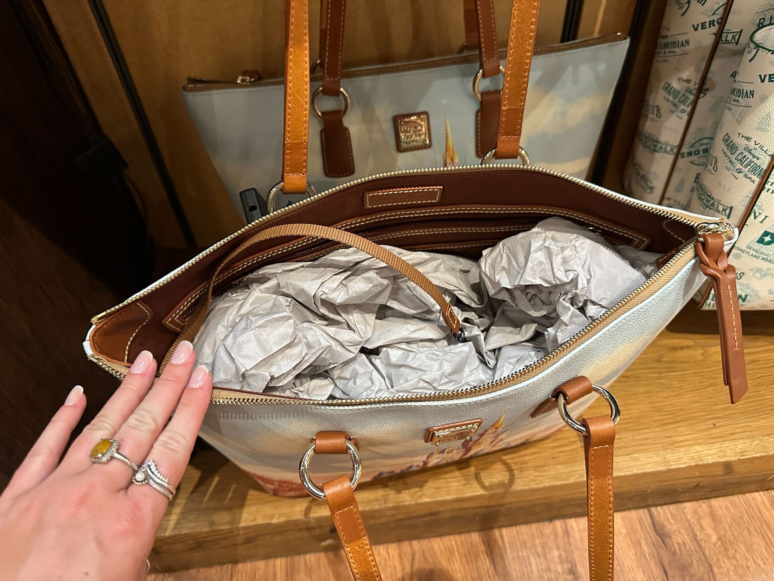 A hand holds open a tan leather handbag filled with crumpled packing paper.