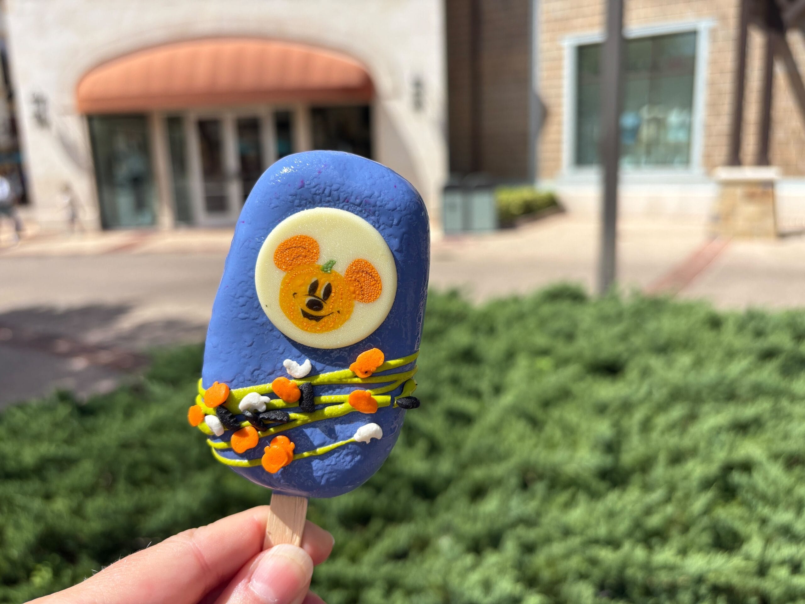 A hand holds a blue Halloween popsicle with a Mickey Mouse pumpkin and icing, spotted at Disney Springs outdoors.