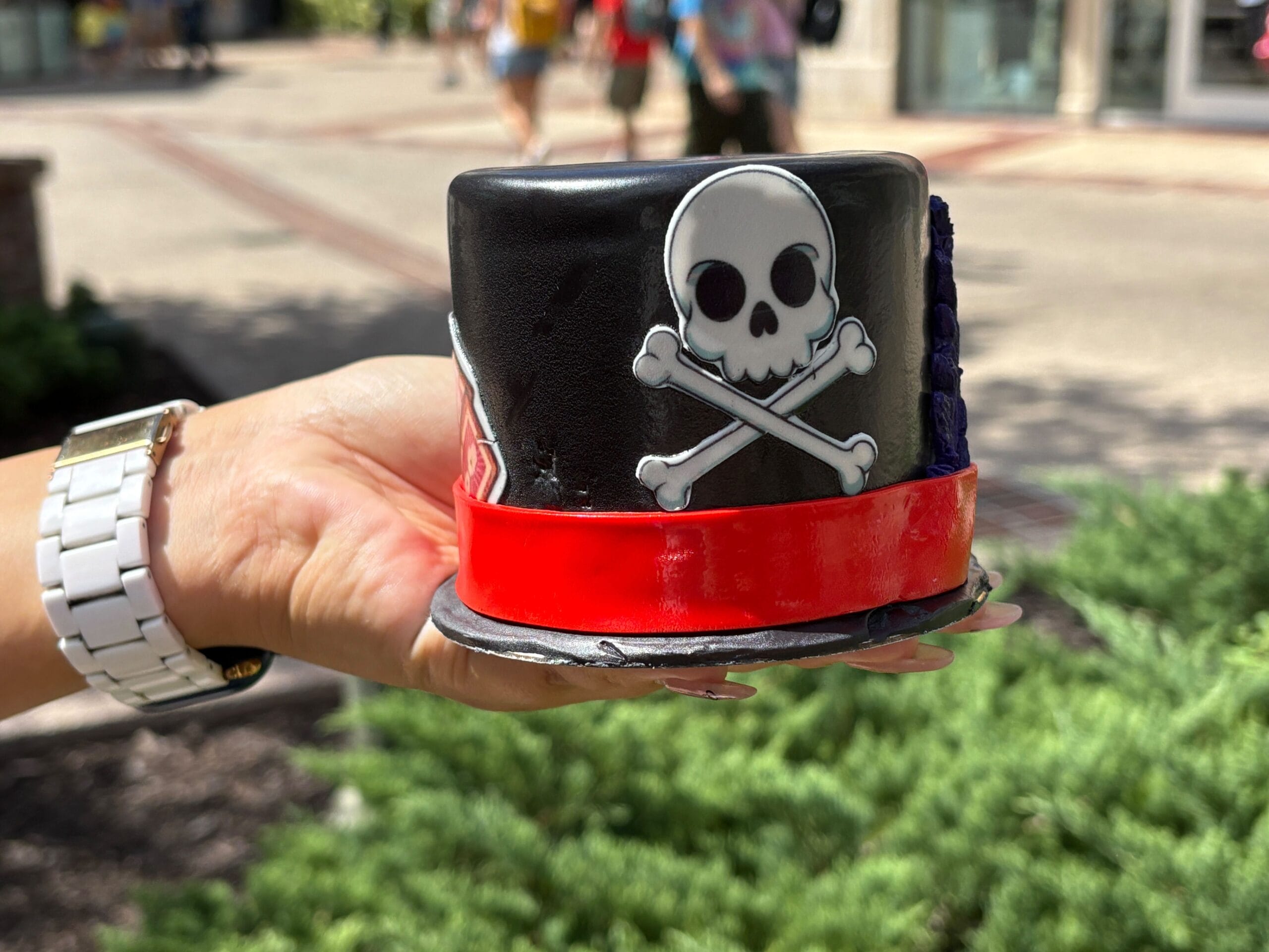 A person holds a small black Halloween food cake with a skull and crossbones design and a red band, outdoors.