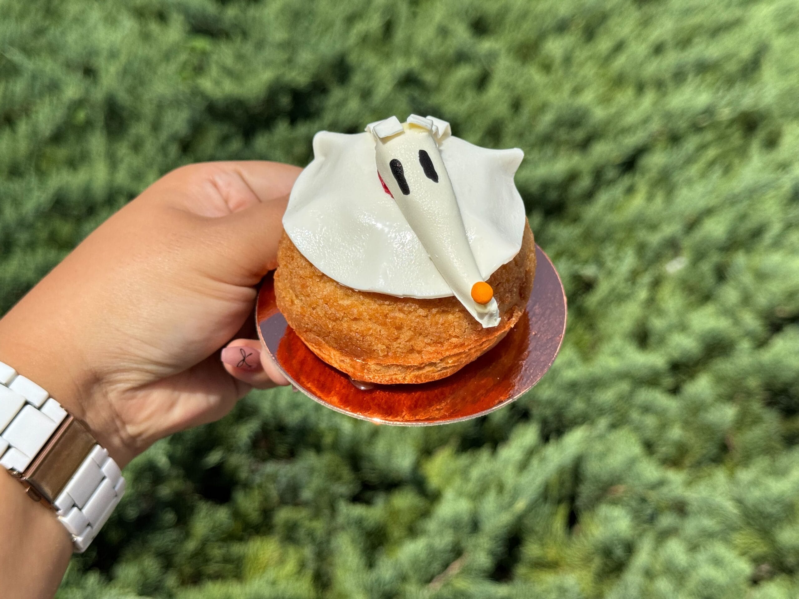 A hand holds a ghost dog pastry, white with an orange nose, at Disney Springs—perfect Halloween food against a green background.