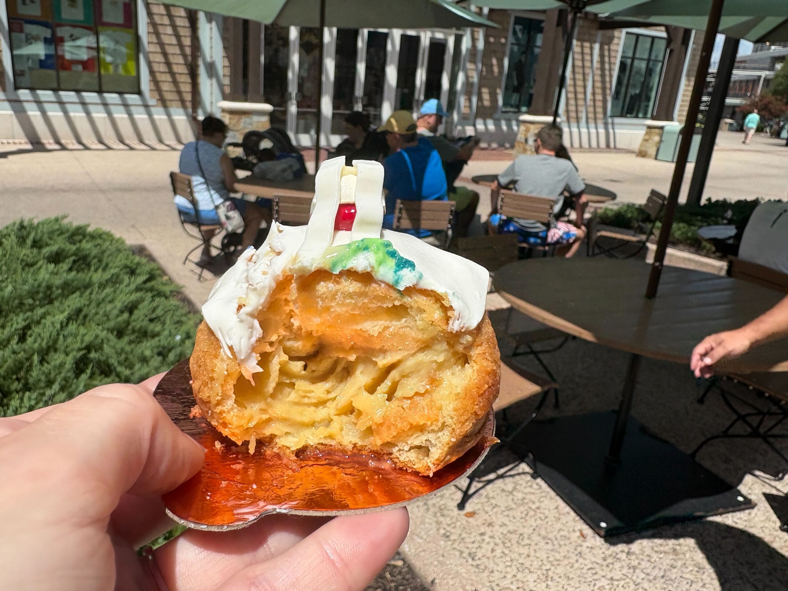 A hand holds a half-eaten pastry with white icing at Disney Springs, outside on a patio with people at tables.