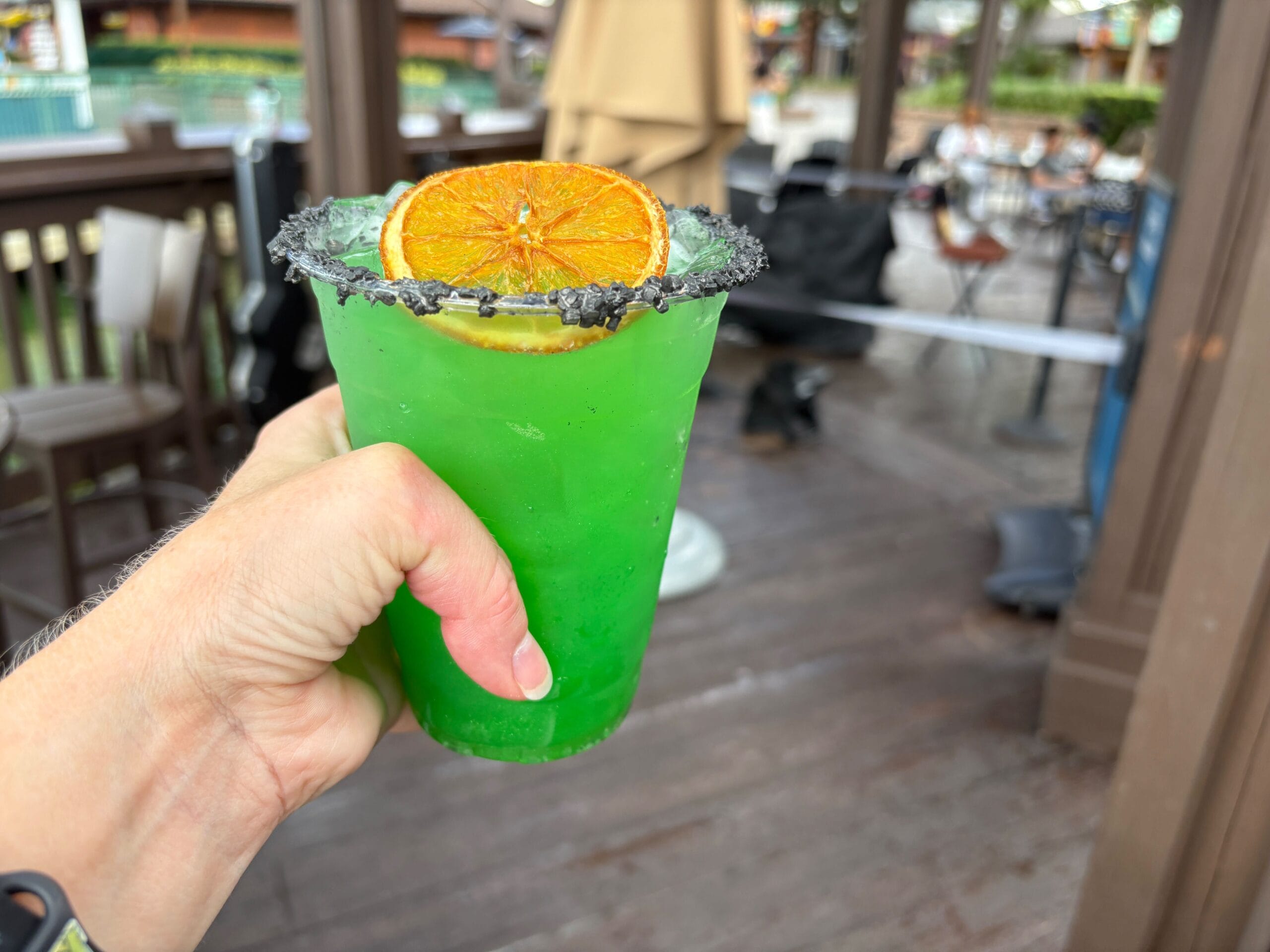 A hand holds a bright green Halloween Food drink with a black salted rim and orange slice garnish outdoors.