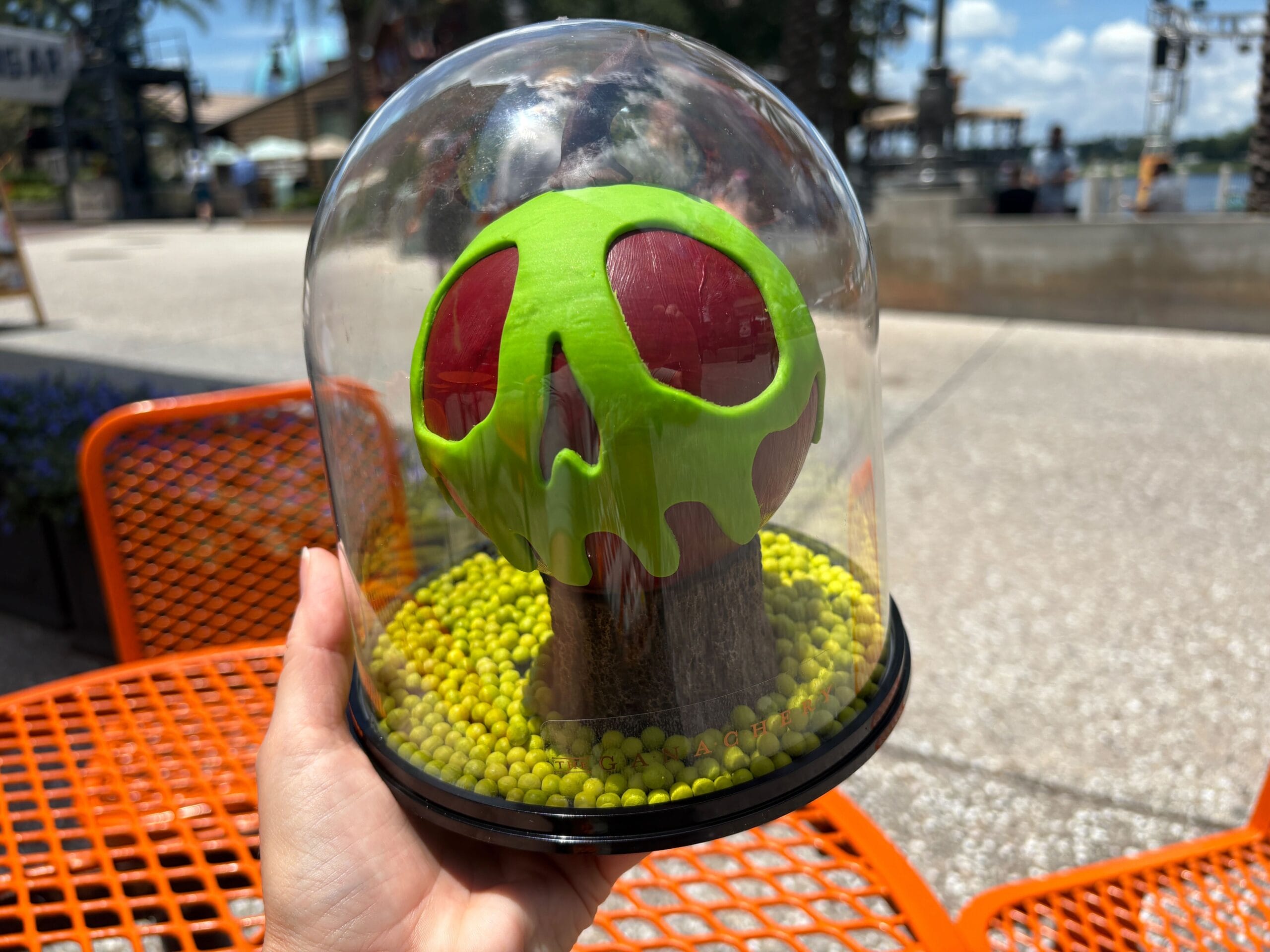 A hand holds a green and red poison apple dome dessert—Halloween food spotted at Disney Springs under a clear sky.