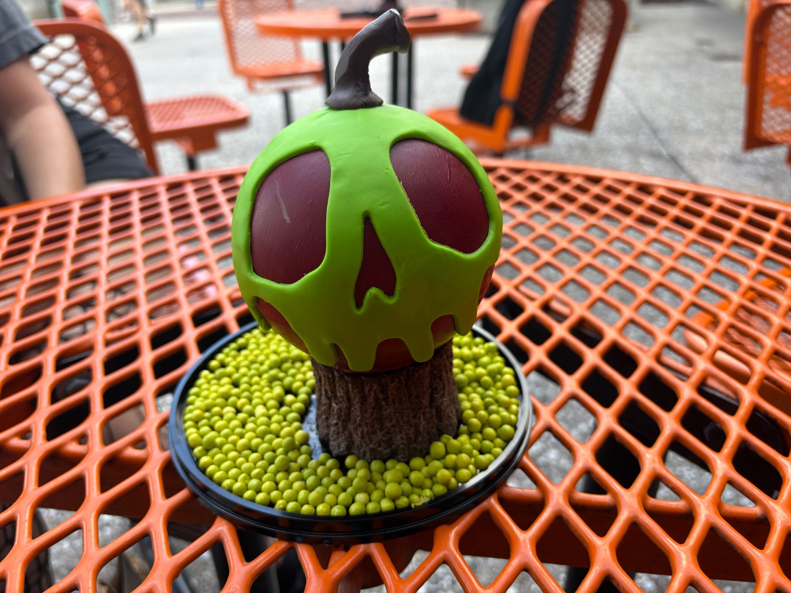 A Halloween food dessert shaped like a green poison apple sits on a black plate with sprinkles, on an orange metal table.