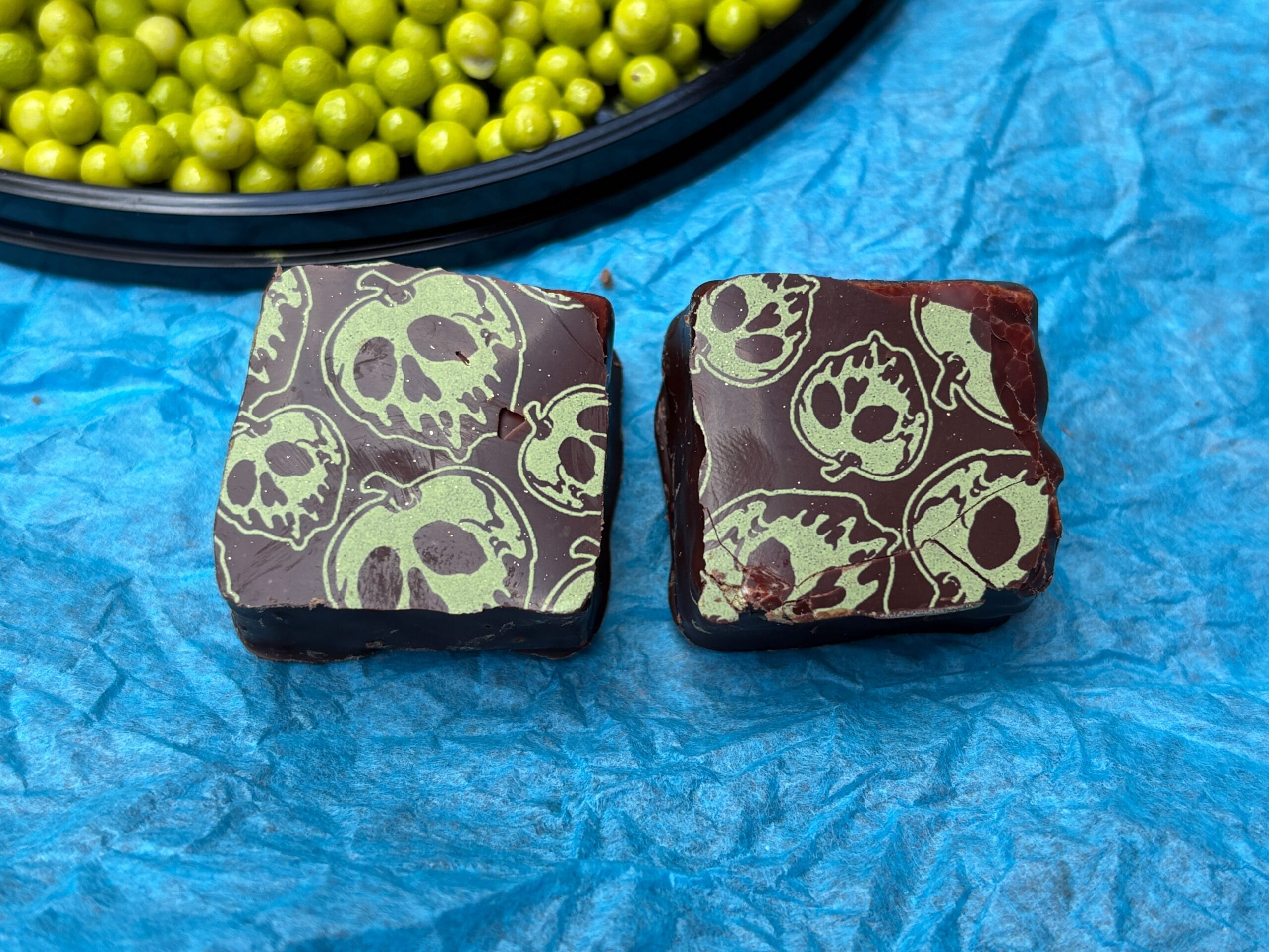 Two chocolate squares with green skulls make spooky Halloween food beside berries on blue fabric.