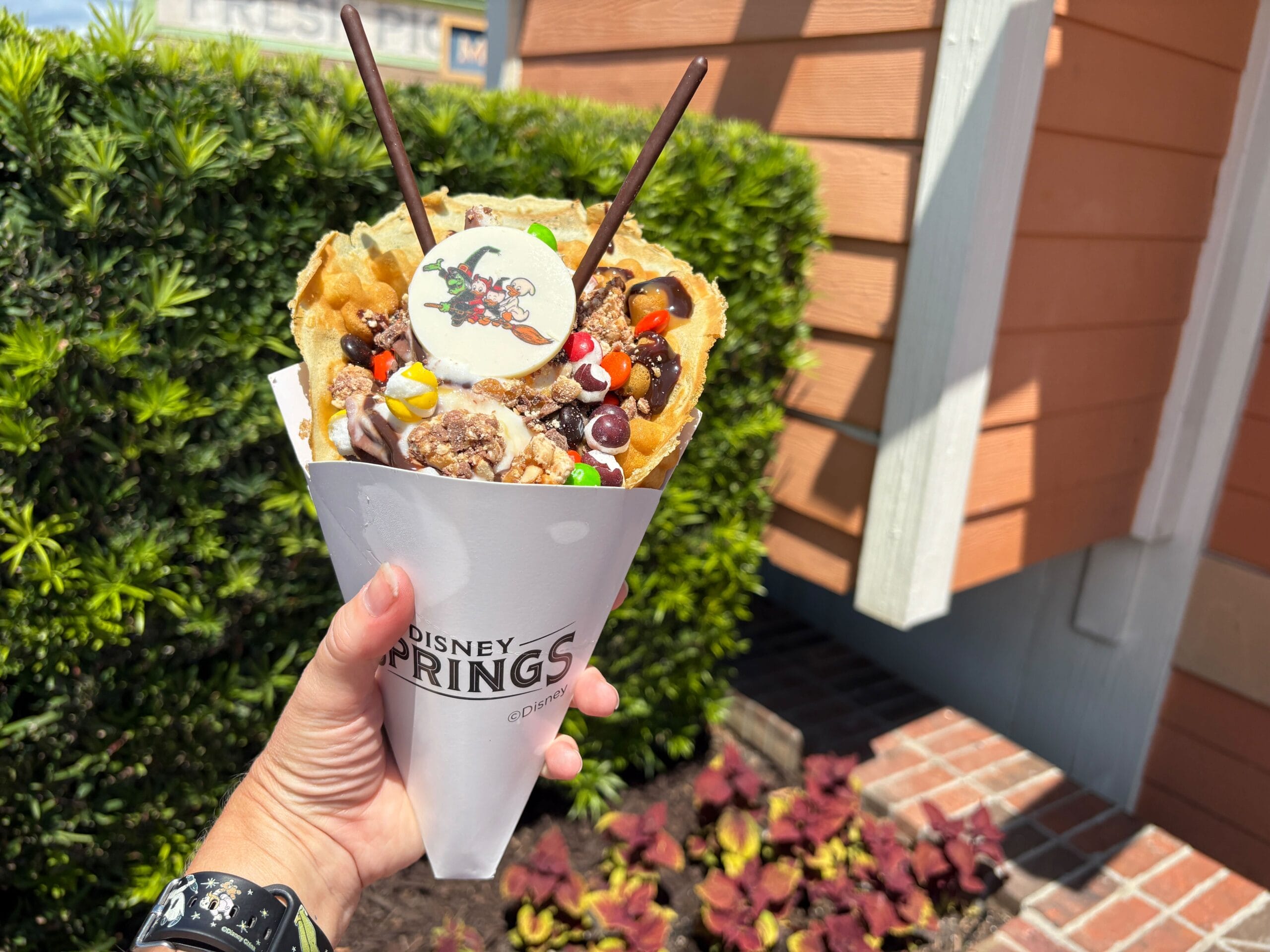 A hand holds a Disney Springs waffle cone topped with Halloween food candy, chocolate sticks, and a printed cartoon wafer.