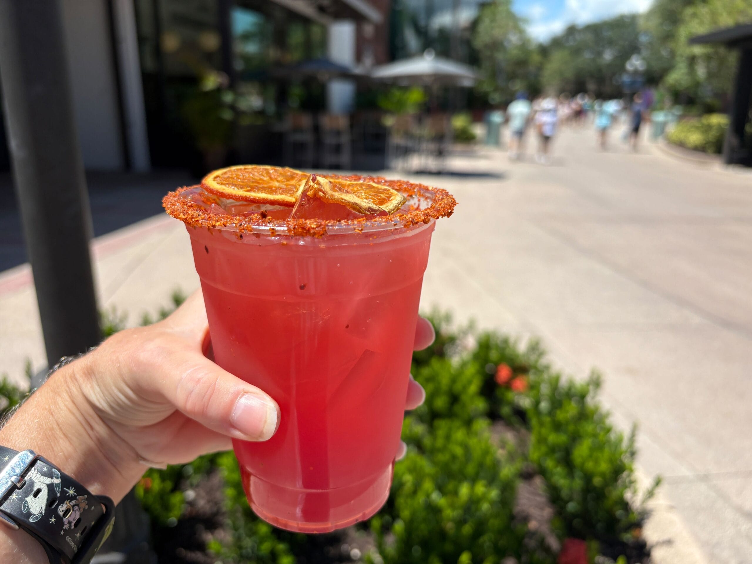 A hand holds a red drink with dried orange slices and chili powder rim at Disney Springs on a sunny day.