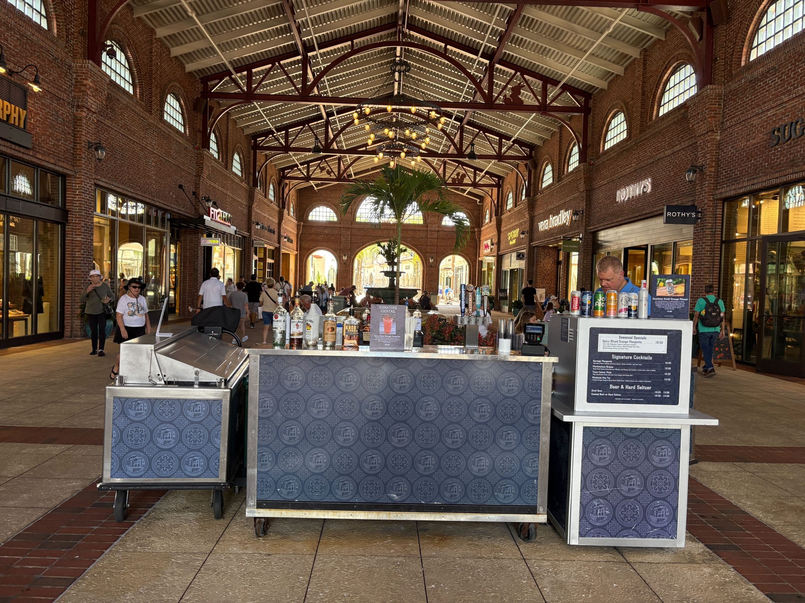 A cocktail bar kiosk in a shopping mall corridor, like those in Disney Springs, with stores and people in the background.