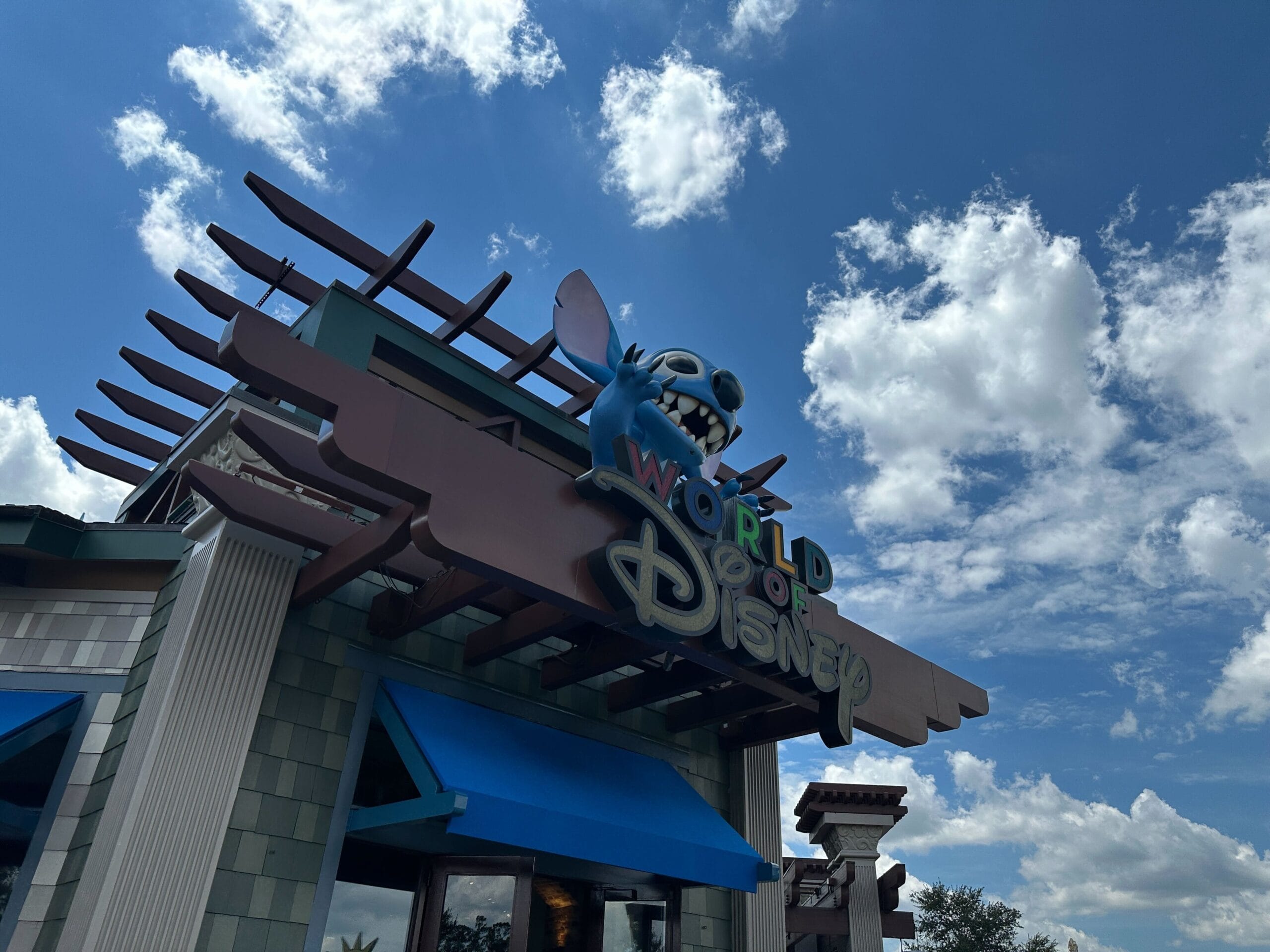 Blue Stitch figure on top of the "World of Disney" store sign against a bright blue sky with clouds.