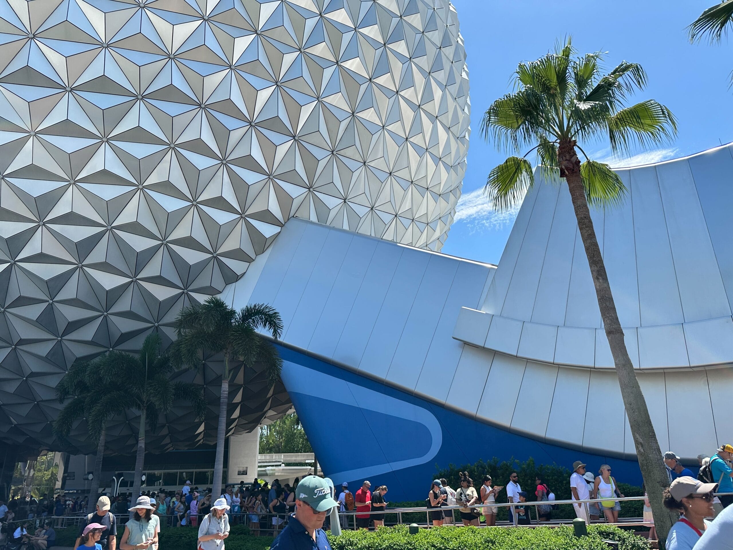 People walking in front of Spaceship Earth at EPCOT, with palm trees and a curved white and blue building in view.
