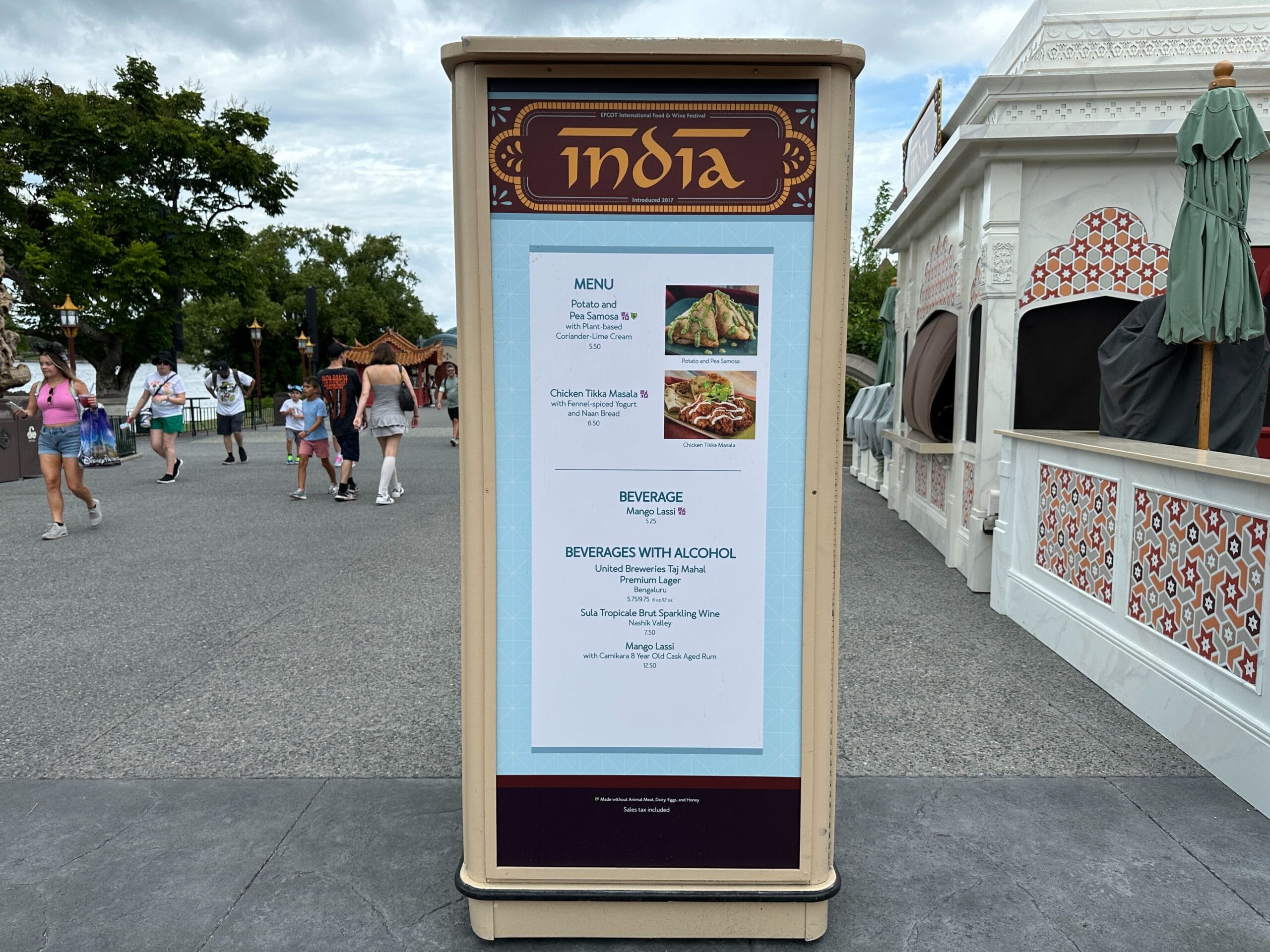 Outdoor menu board at EPCOT's India pavilion for the 2025 Food & Wine Festival, with people strolling nearby.
